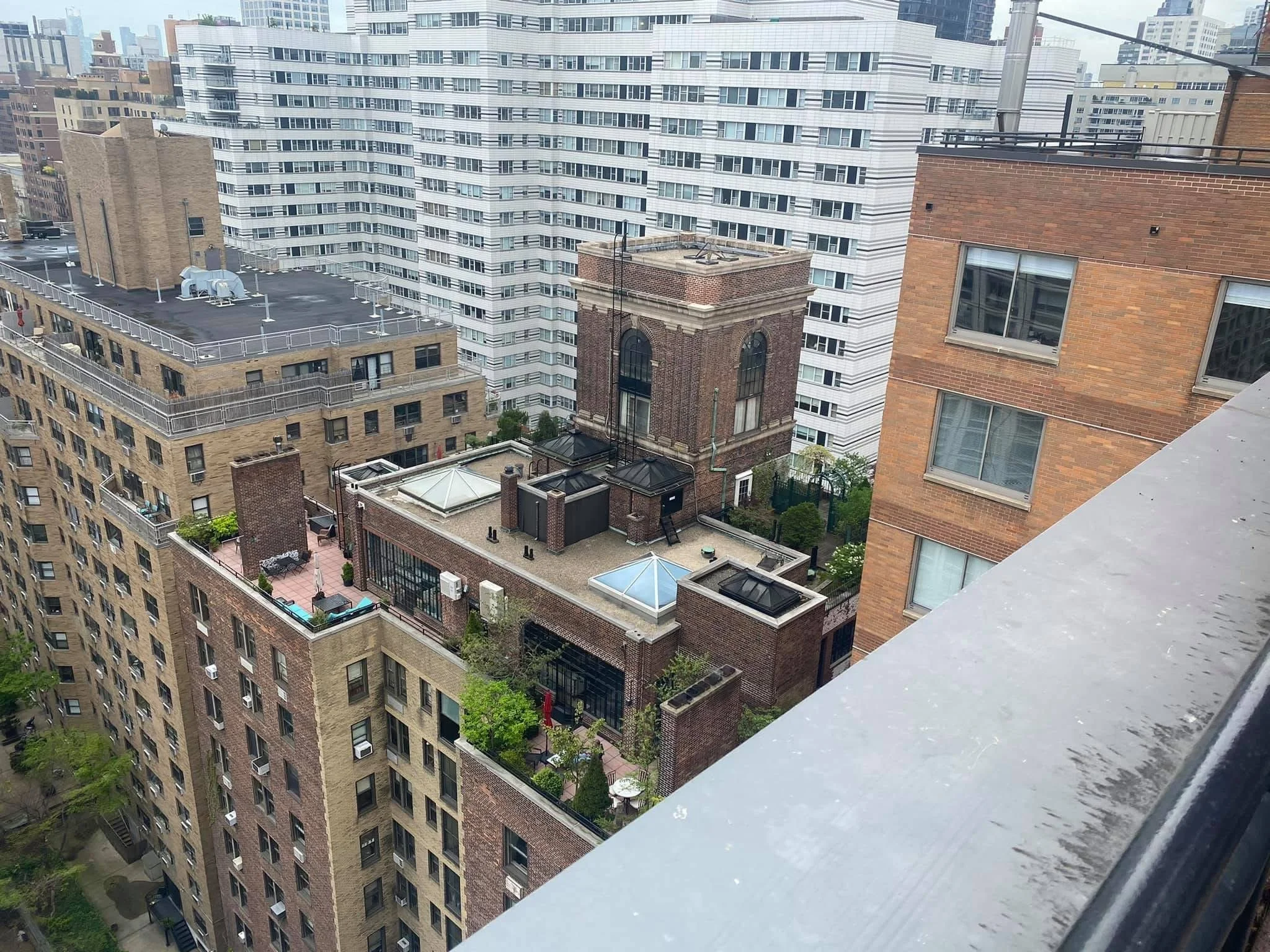 Cityscape view from a high-rise balcony showing multiple residential buildings with rooftop terraces, windows, and urban streets below.