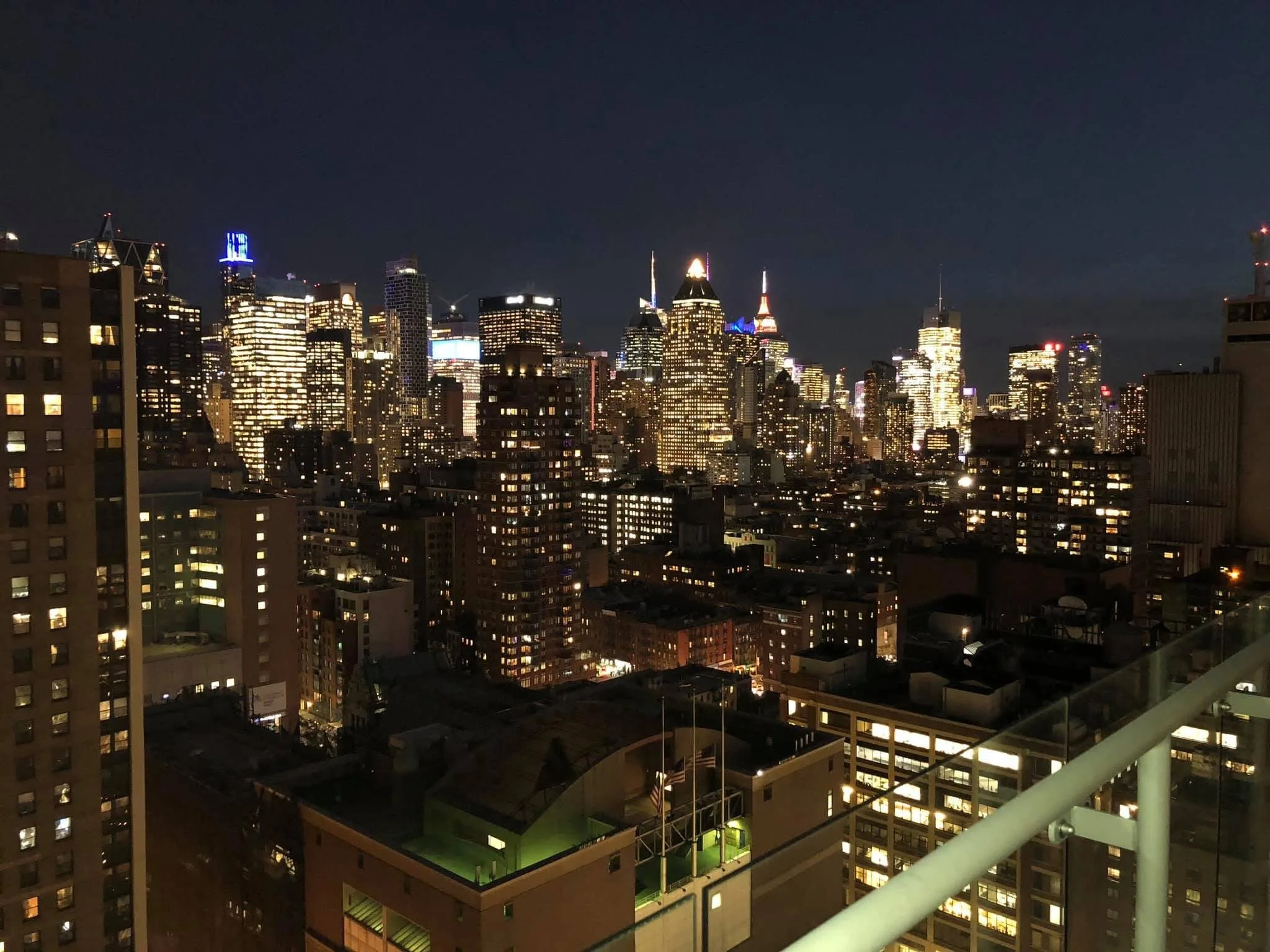 Nighttime city skyline view of a densely built urban area with high-rise buildings, some illuminated, under a dark sky, taken from a balcony.