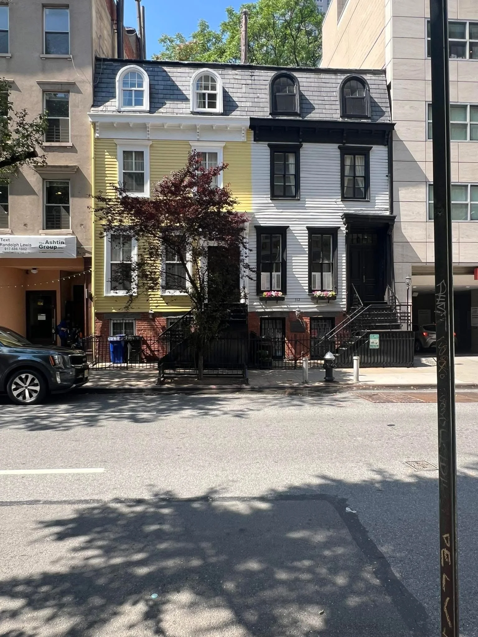 Three-story house with a distinctive black-and-white color scheme, featuring a yellow section on the left side. The house has black stairs and railing leading up to a black front door, with flower boxes on the windows. There is a small tree in front 