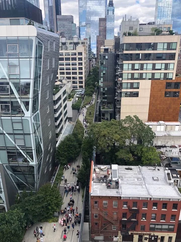 A cityscape view from a tall building, showing a busy pedestrian street with people walking along it, flanked by modern and older buildings, with skyscrapers in the background.
