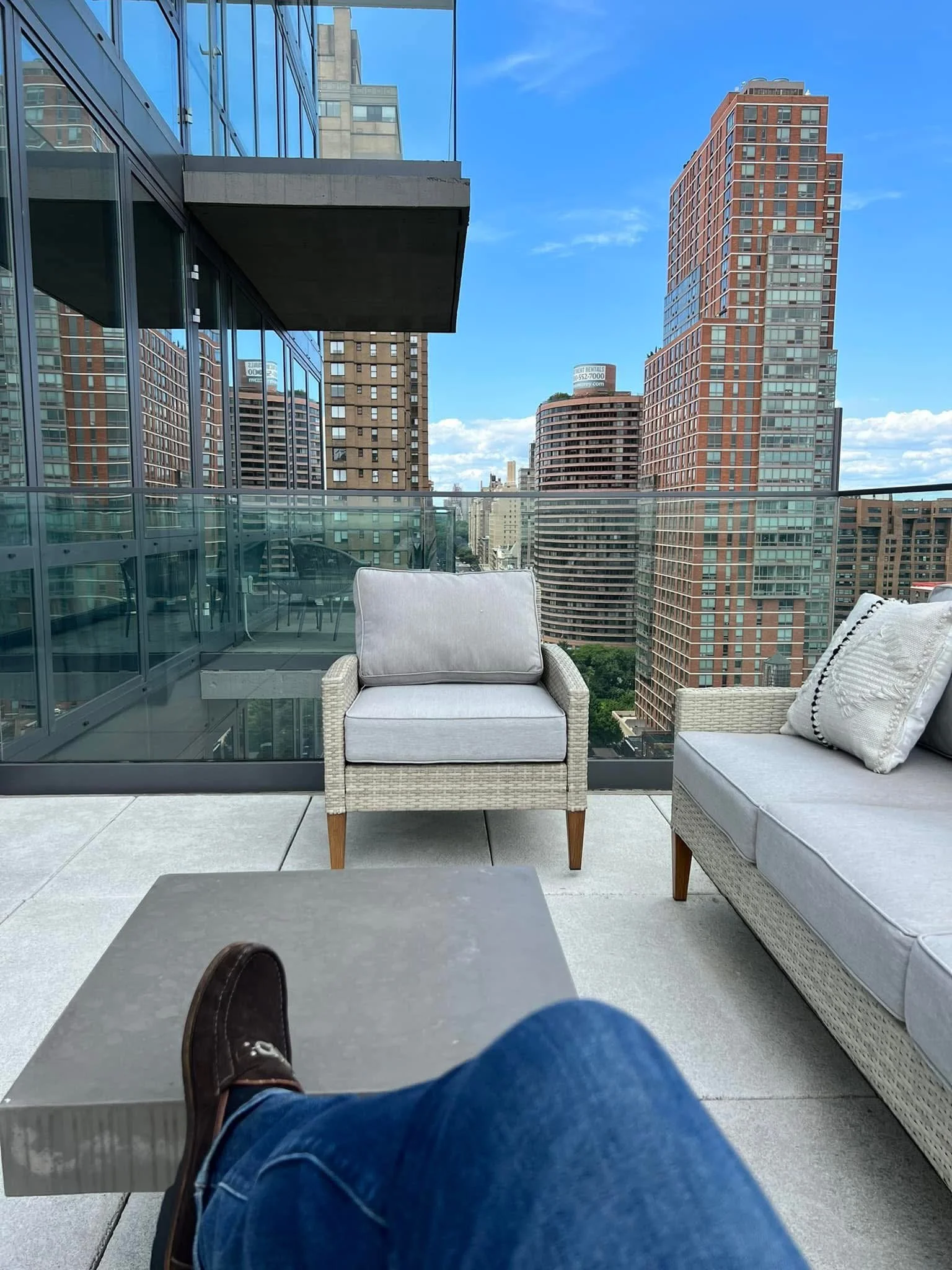 A balcony with outdoor furniture including a white cushioned armchair and a sofa. The person taking the photo is seated, with their foot resting on a table, wearing jeans and brown shoes. The balcony has glass railings and offers a view of tall city buildings under a blue sky with some clouds.