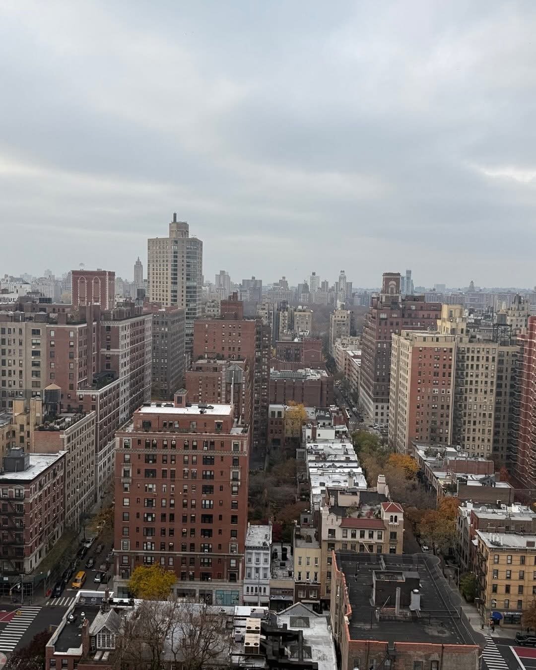 Overcast cityscape of New York City with tall buildings, some with rooftop features, and streets below, showing cars and trees with autumn foliage.