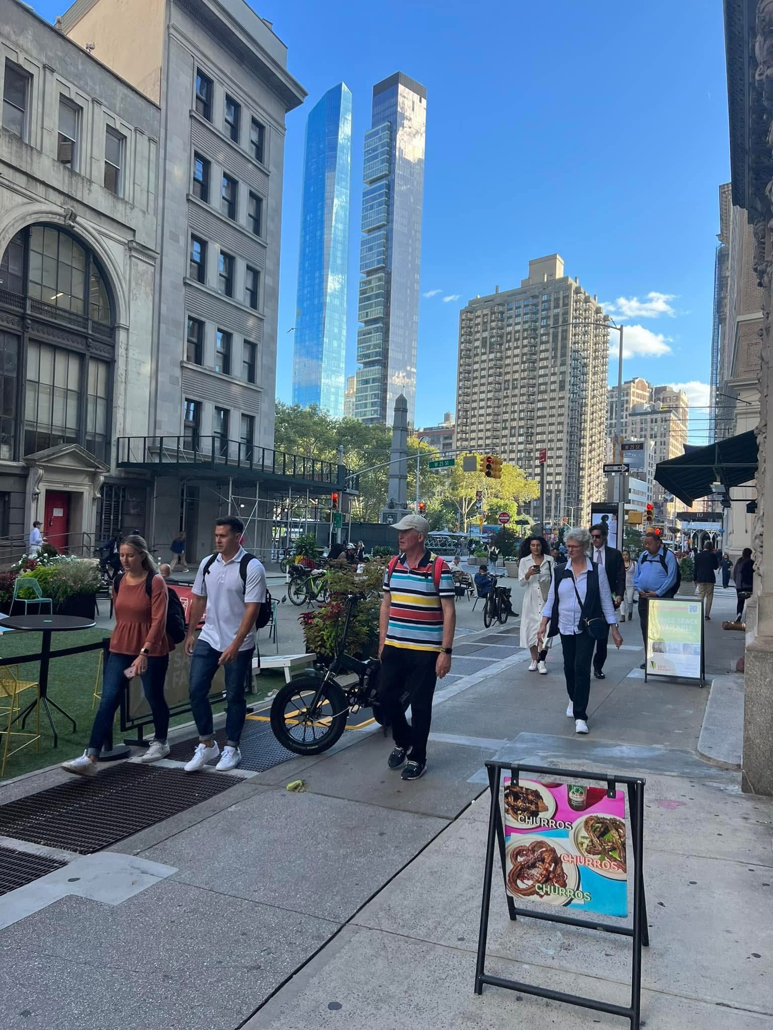 City street scene with pedestrians walking on the sidewalk, modern skyscrapers in the background, and a sign advertising churros in the foreground.