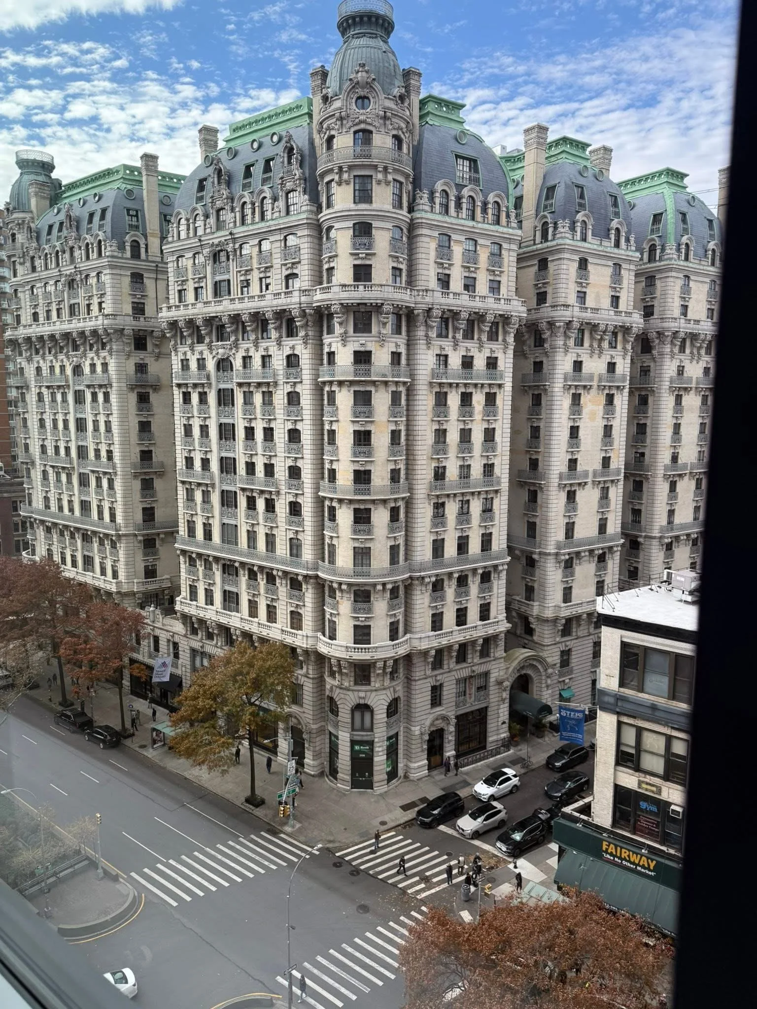 A large, historic-style apartment building with ornate architectural details, multiple balconies, and a domed rooftop, situated on a city street with parked cars and pedestrians, under a partly cloudy sky.