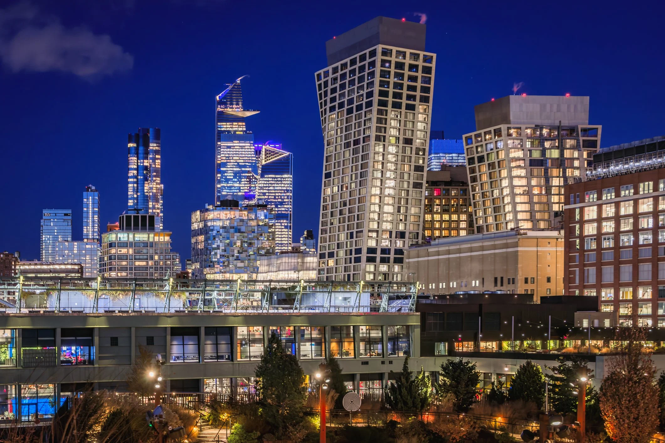 Nighttime cityscape of a modern downtown with illuminated skyscrapers, a rooftop terrace with lights, and trees in the foreground.