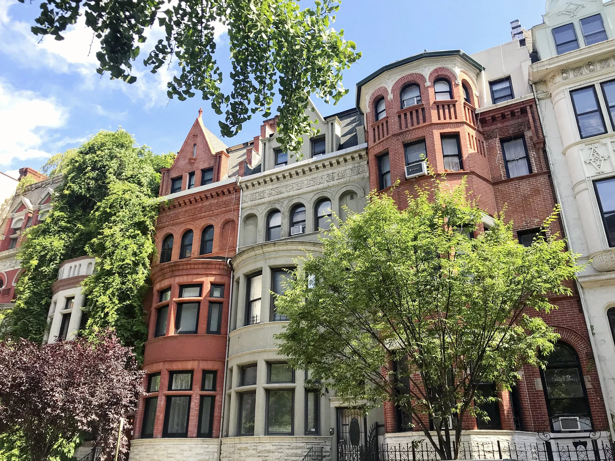 Street view of historic brownstone and brick row houses with trees in front, under a partly cloudy sky.