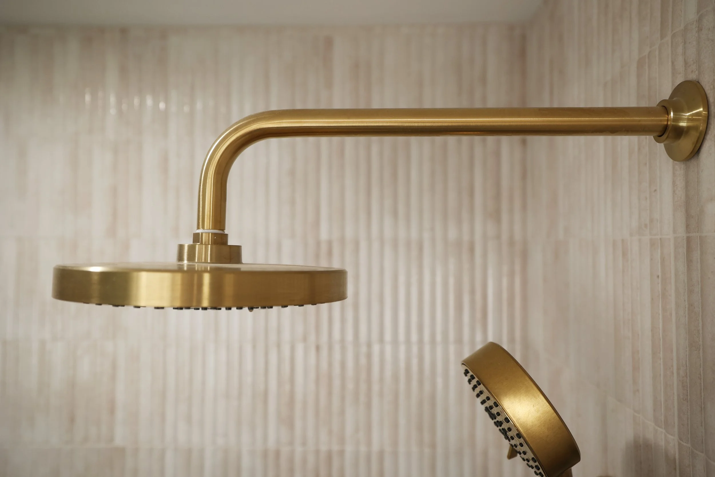 Brass rain shower head mounted on vertically fluted tile in a warm organic modern bathroom.