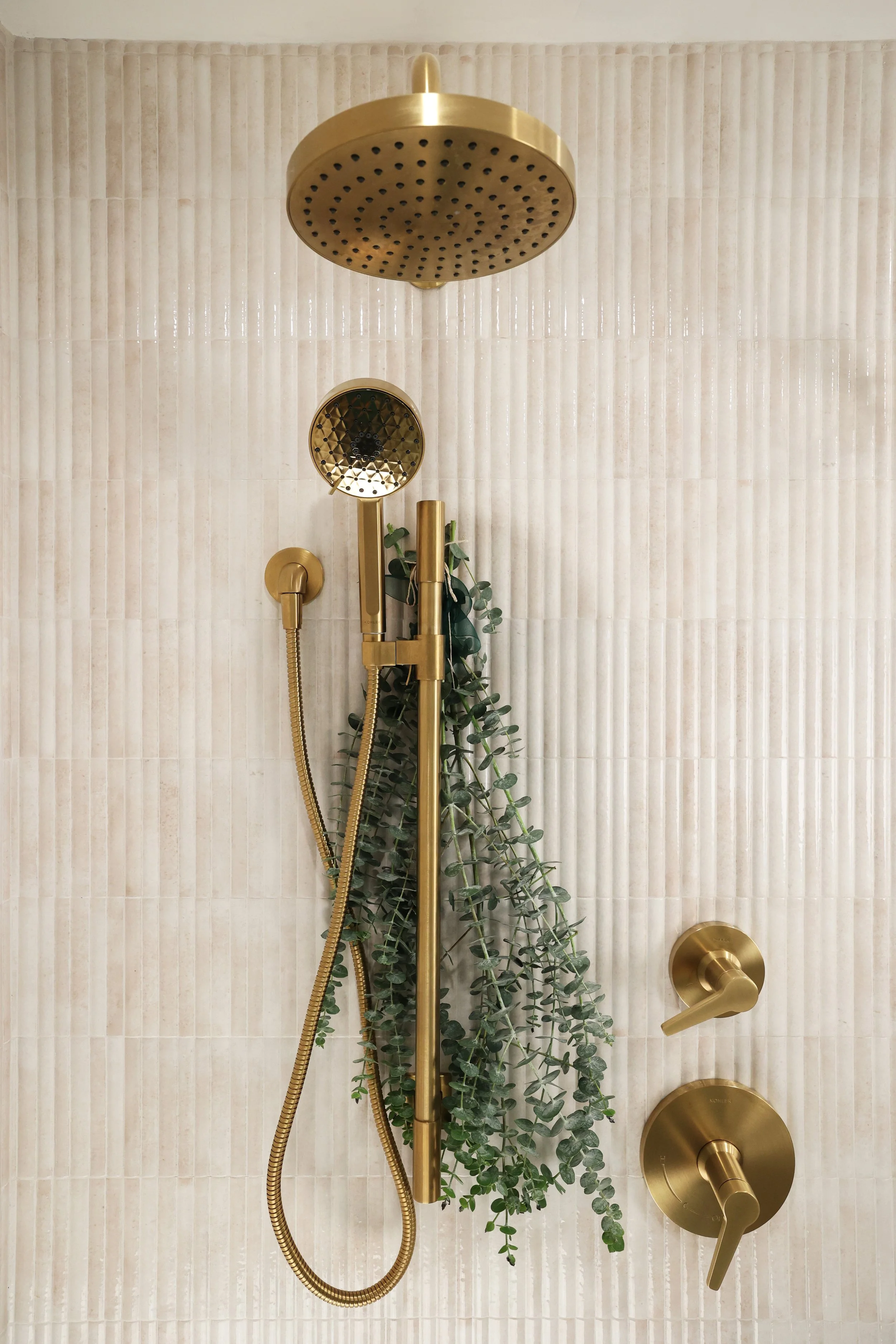 Polished brass shower system against fluted neutral tile in an organic modern primary bathroom.