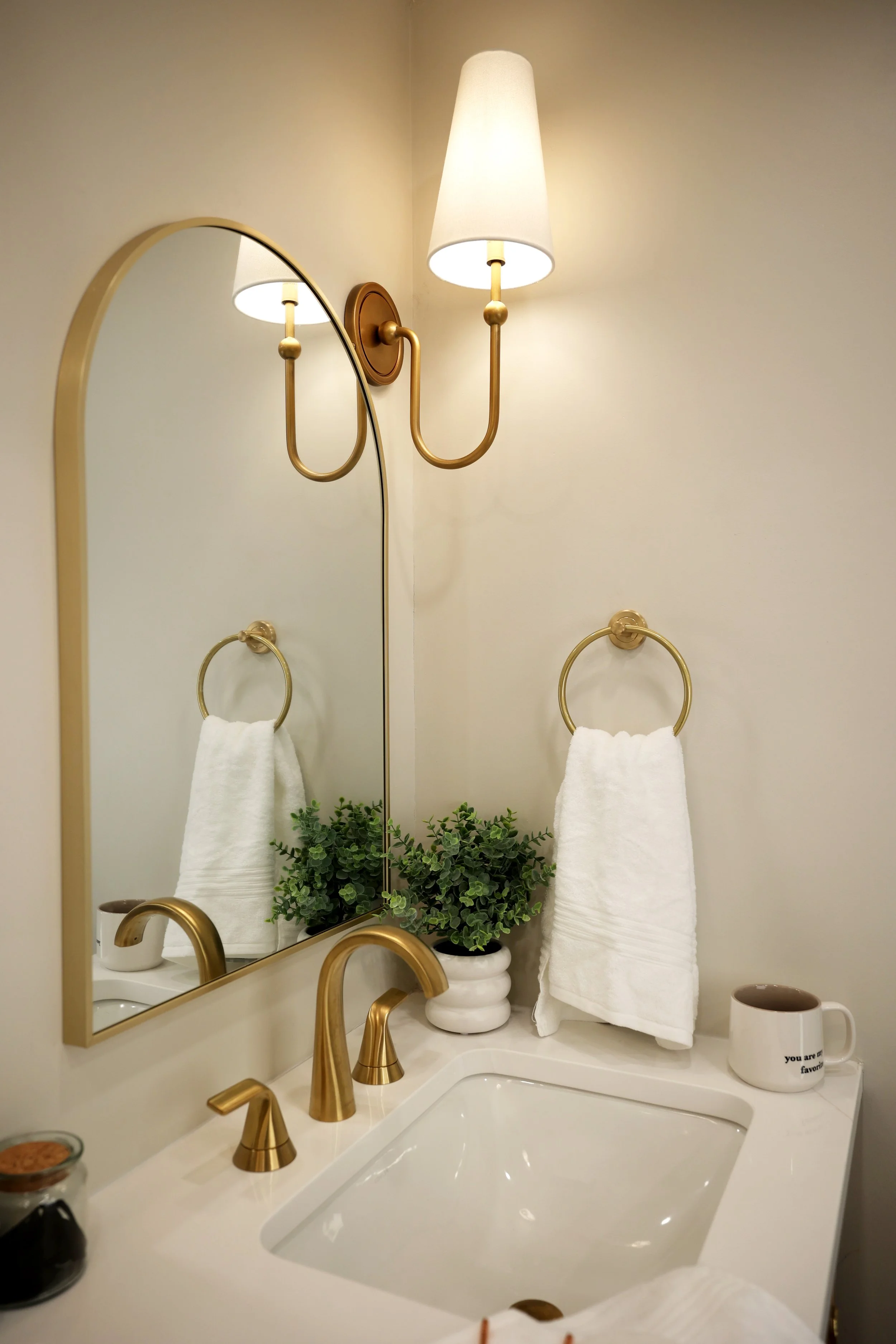 Arched mirror and brass wall sconce above oak vanity in a soft, organic modern primary bathroom.