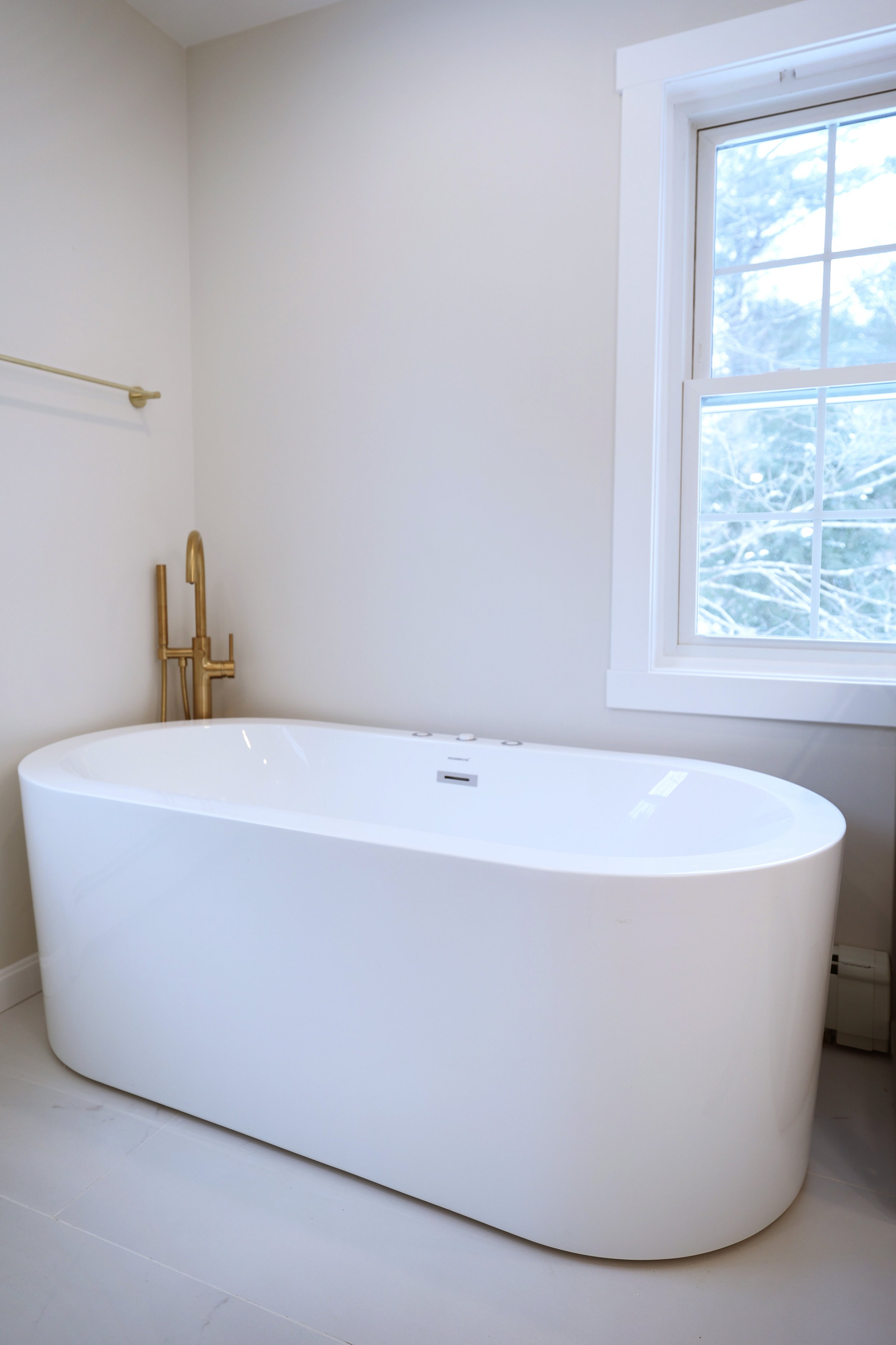 Freestanding soaking tub beneath a window in a marble and brass bathroom in Lebanon New Hampshire.