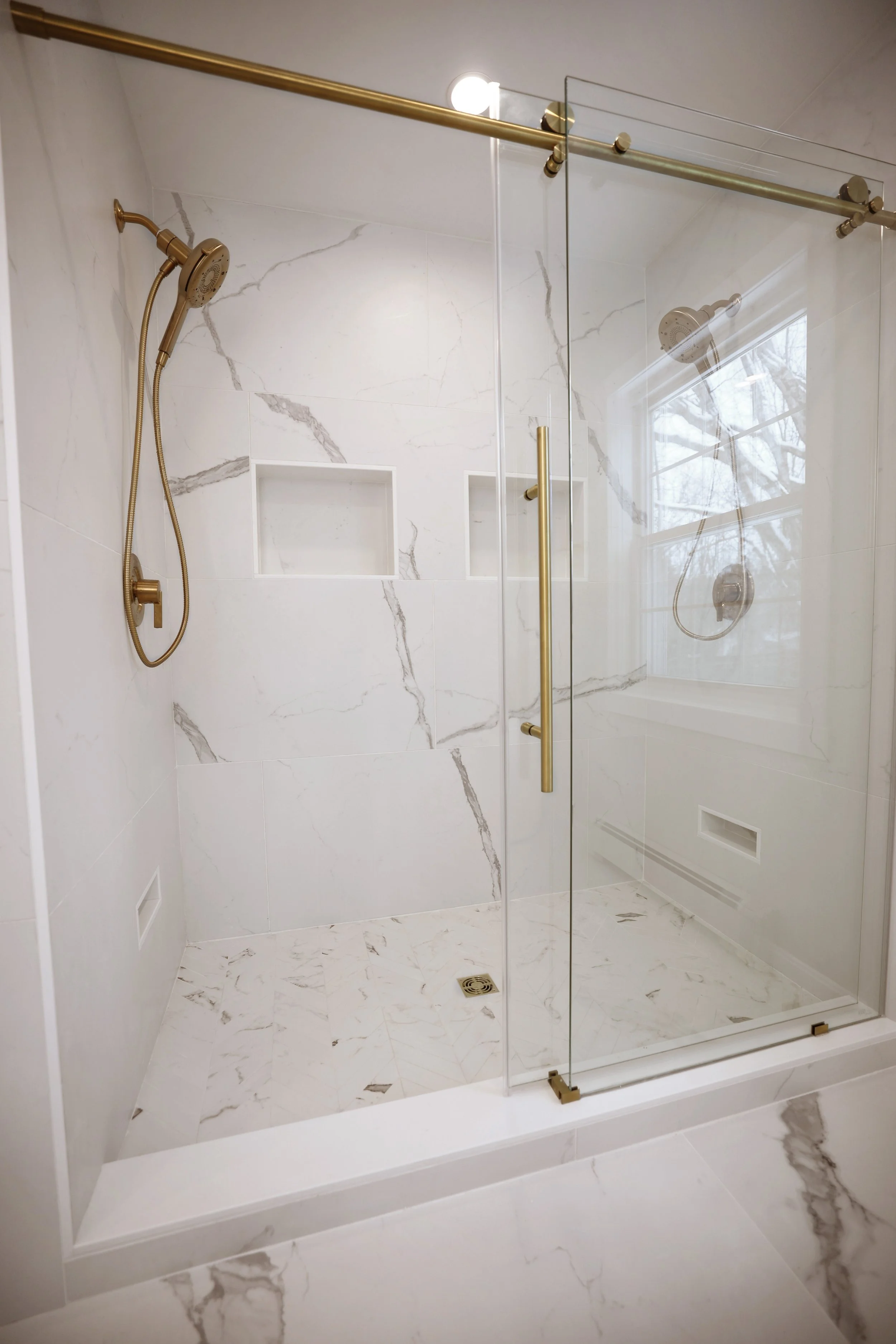 Glass shower with marble-look tile and brass Moen Cia fixtures in a primary bathroom renovation in Lebanon NH.