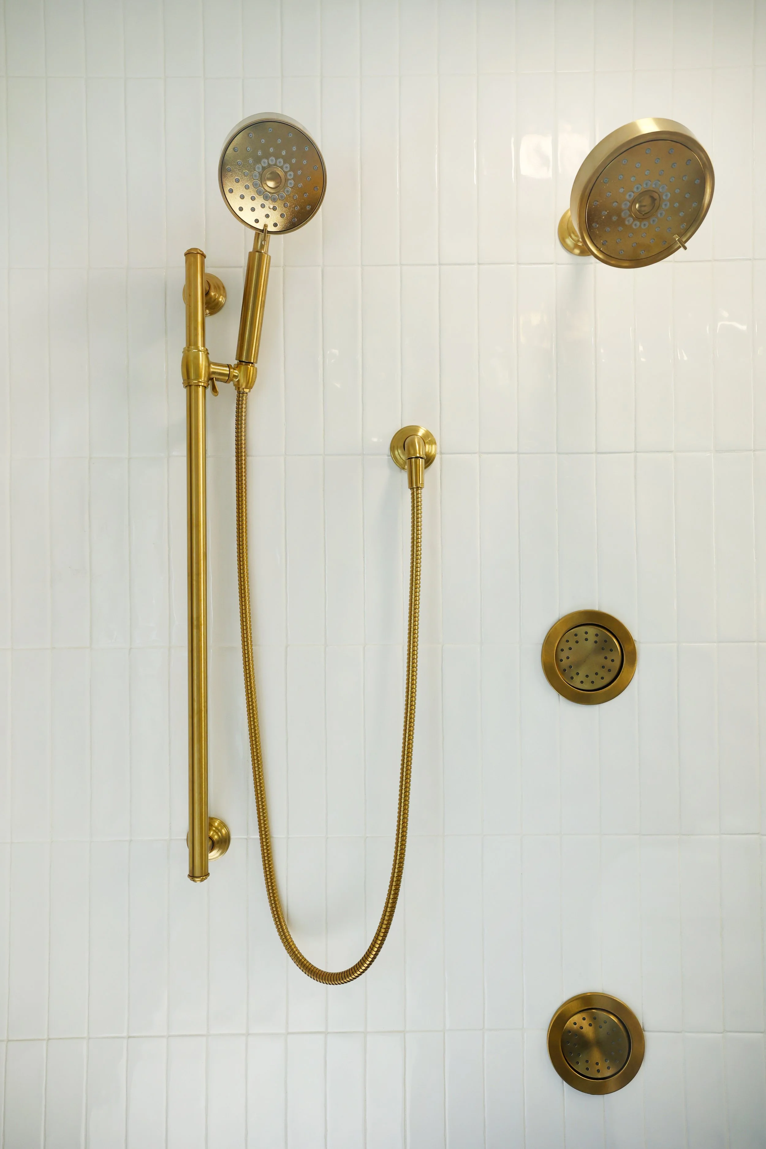 Brass shower system with handheld fixture set against white tile in a black and brass bathroom.