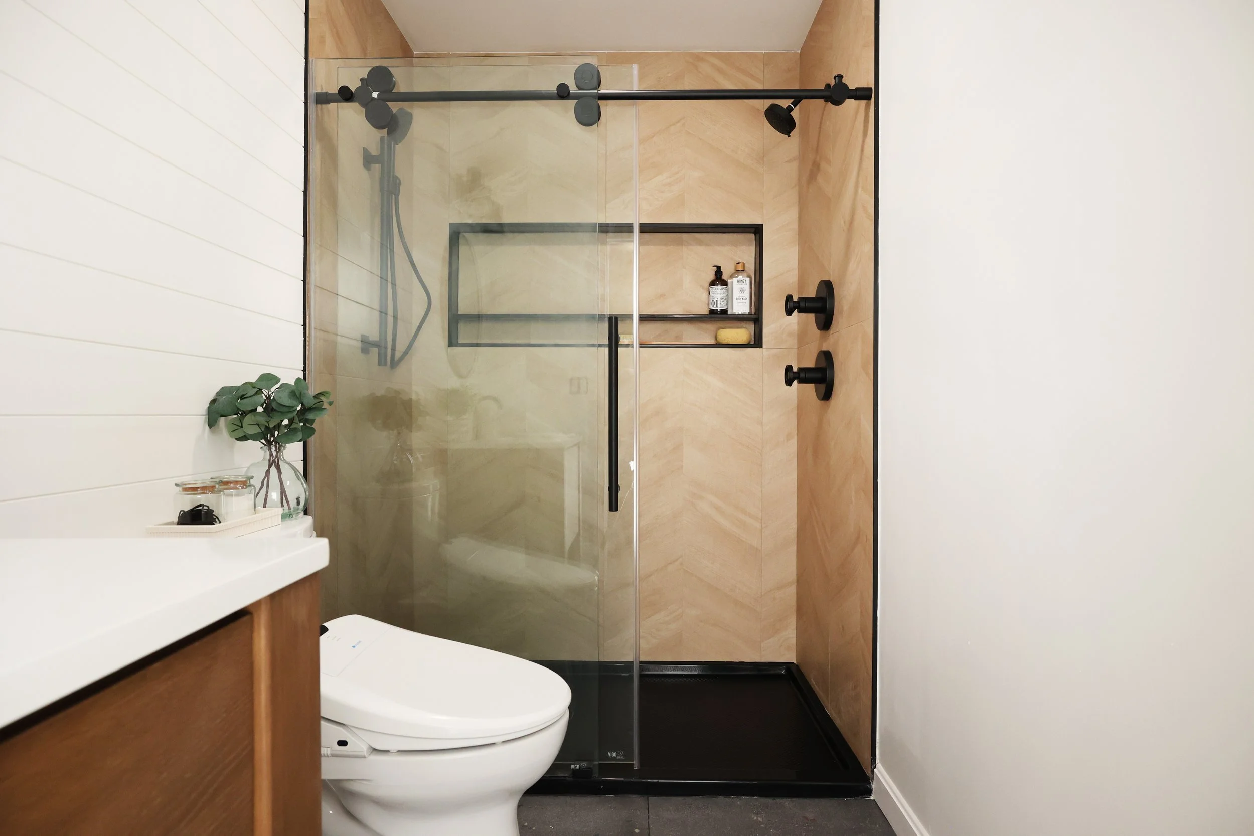  Walk-in shower with wood look chevron tile, matte black fixtures, glass enclosure, and built-in shower niche in a modern bathroom renovation. 