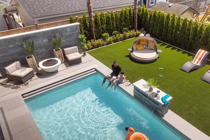 A backyard pool area with two people sitting on the pool's edge, a man and a girl. There is greenery and outdoor furniture, including a circular daybed, two lounge chairs, and two armchairs with pillows. The yard is enclosed by tall hedge fences.