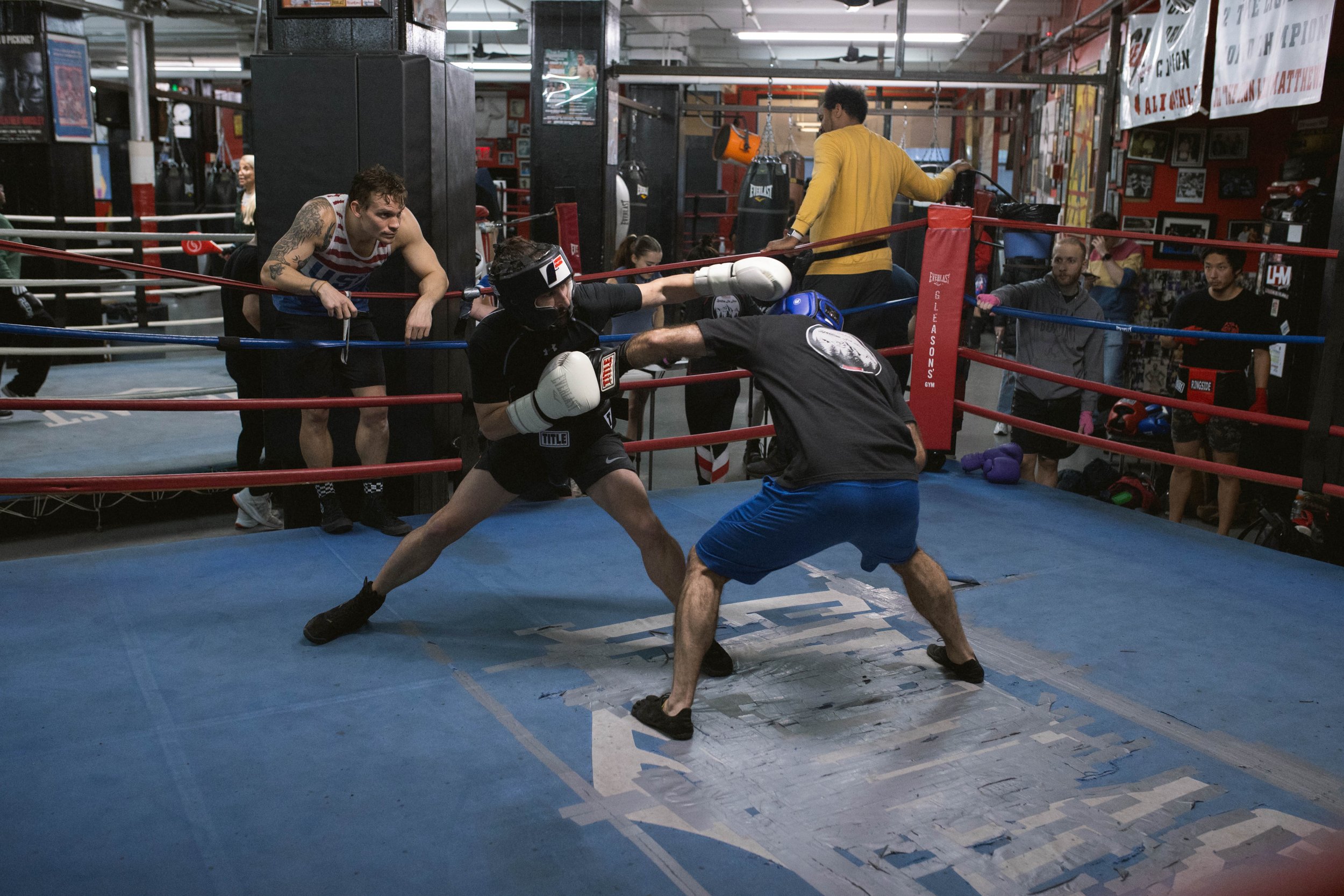 Boxers sparring in a gym ring, observed by trainers and other boxers.