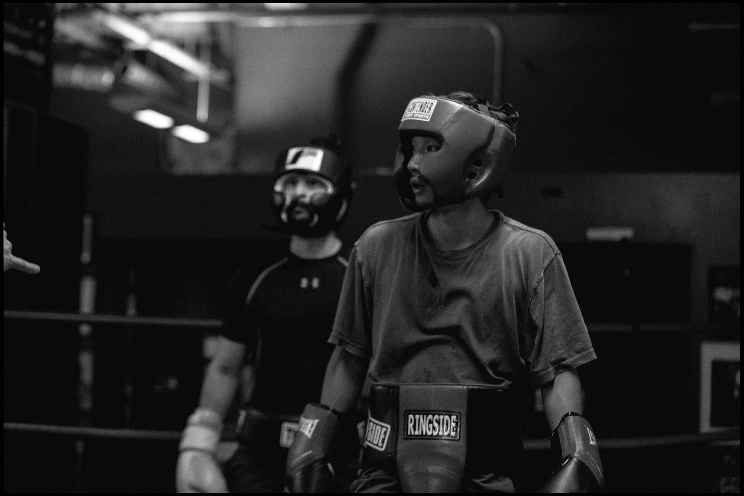Two people wearing headgear and gloves prepare for boxing in a dimly lit gym.