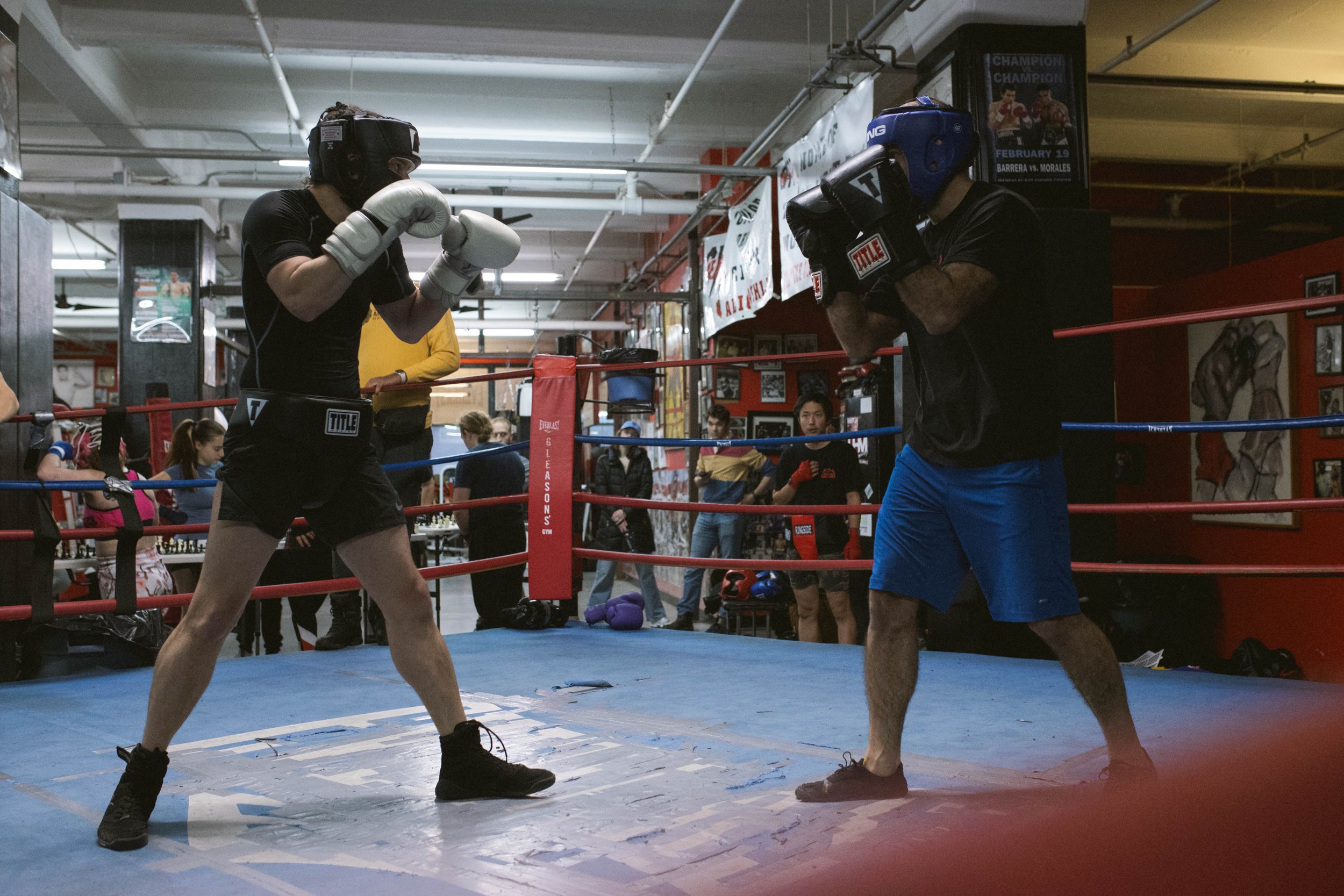 Two people wearing boxing gear sparring in a boxing ring with spectators nearby.