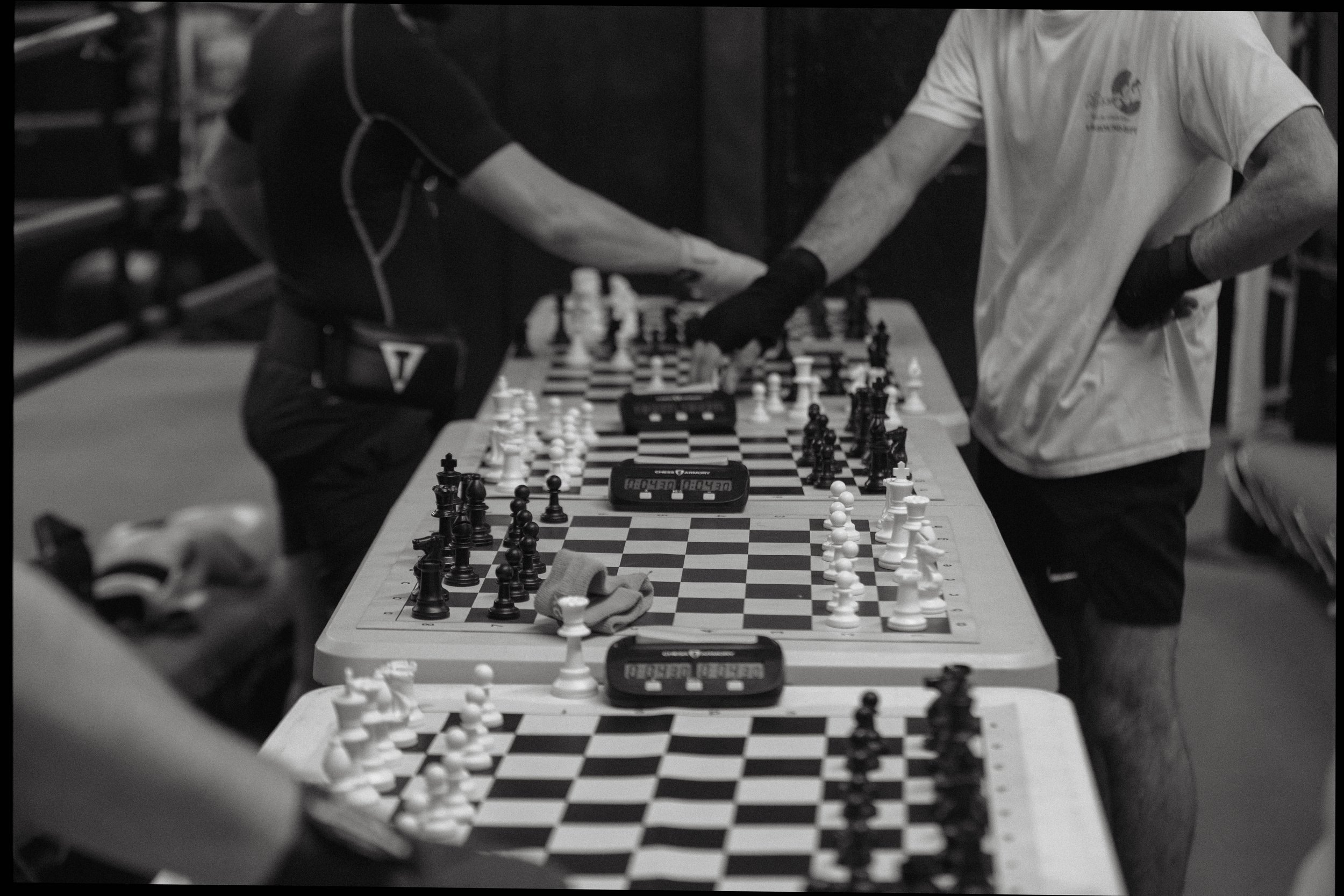 Black and white photo of two people shaking hands over a chessboard, with multiple chessboards set up on tables, and a chess clock visible. Both individuals are wearing casual sportswear and gloves.