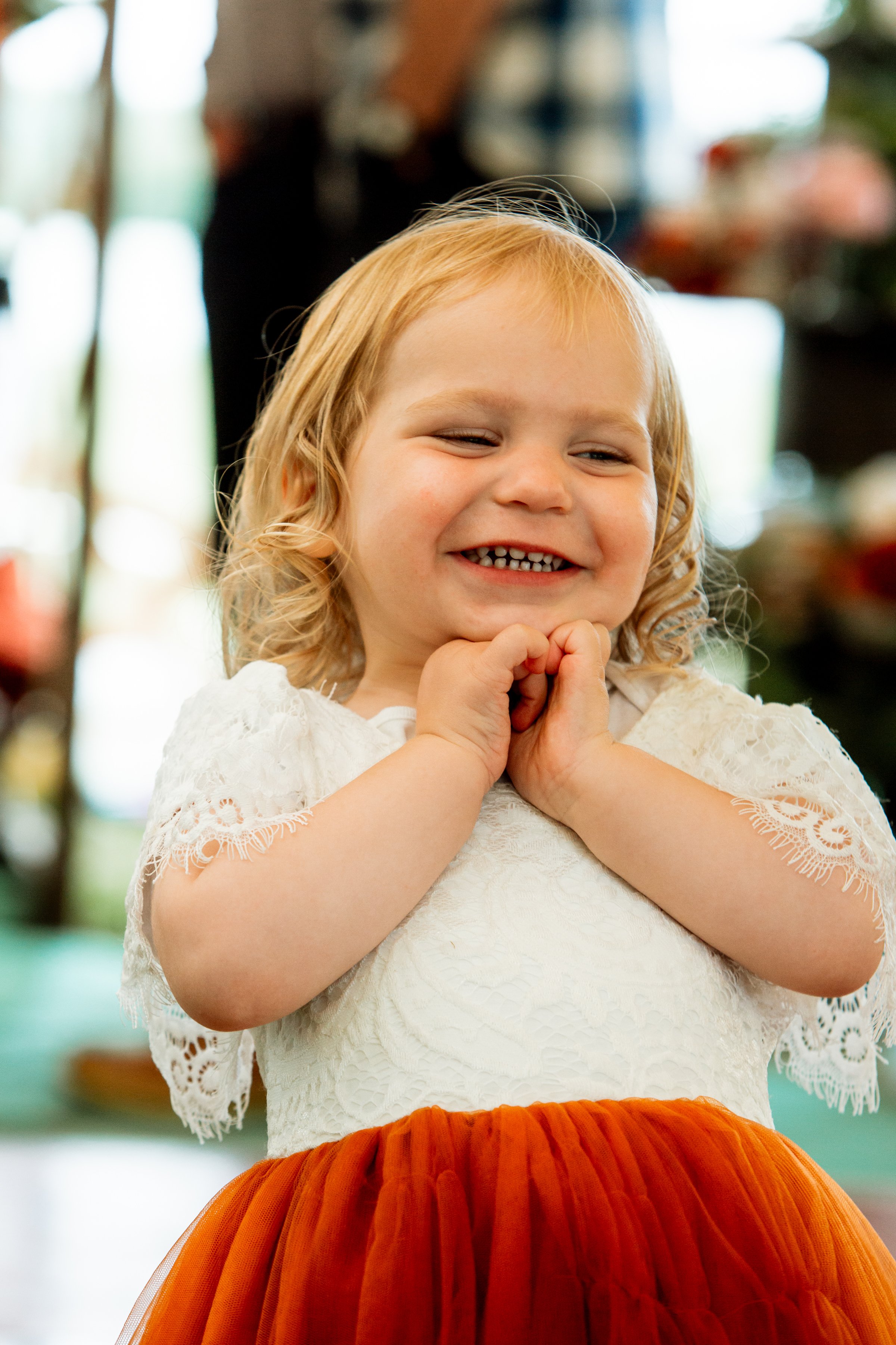A little girl in a white lace dress smiles with delight, holding her hands beneath her chin during a wedding celebration.