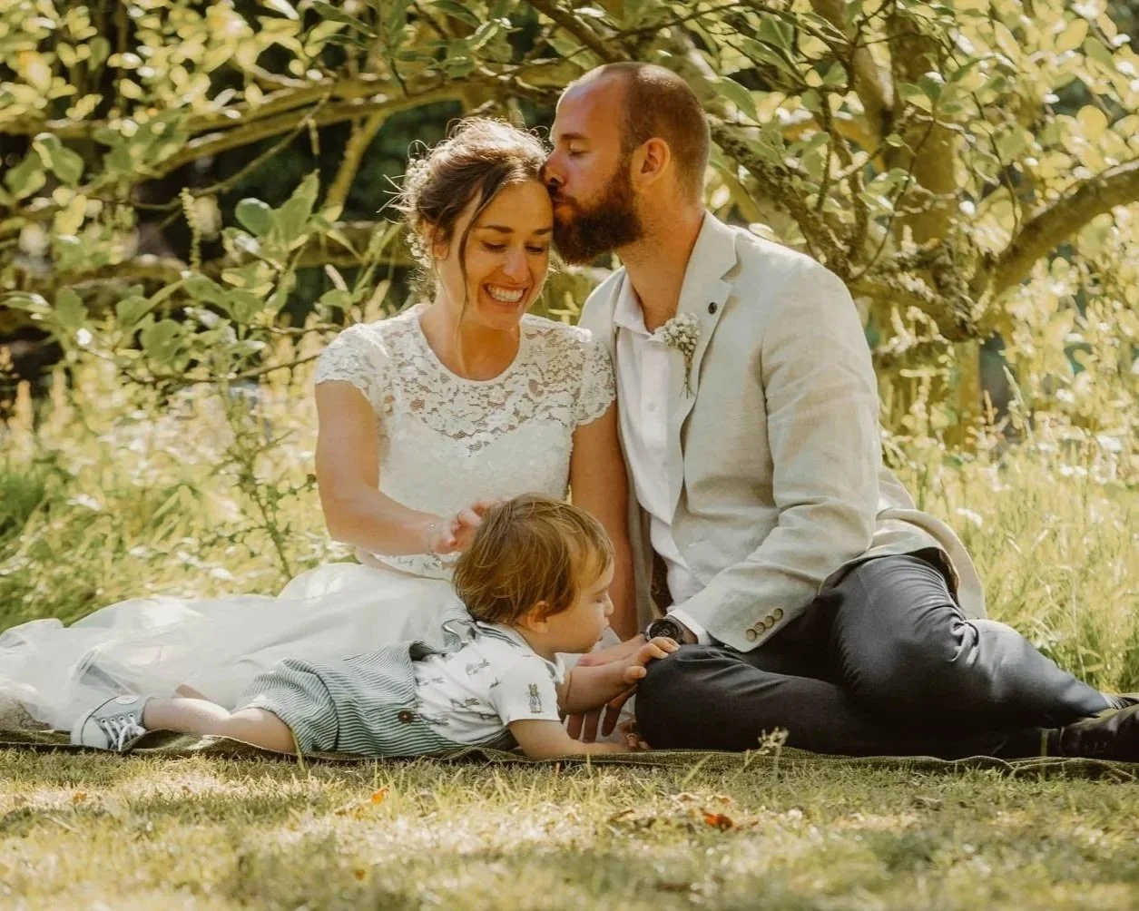 A groom kisses his bride on the forehead as their young child plays beside them on the grass during an intimate outdoor wedding.