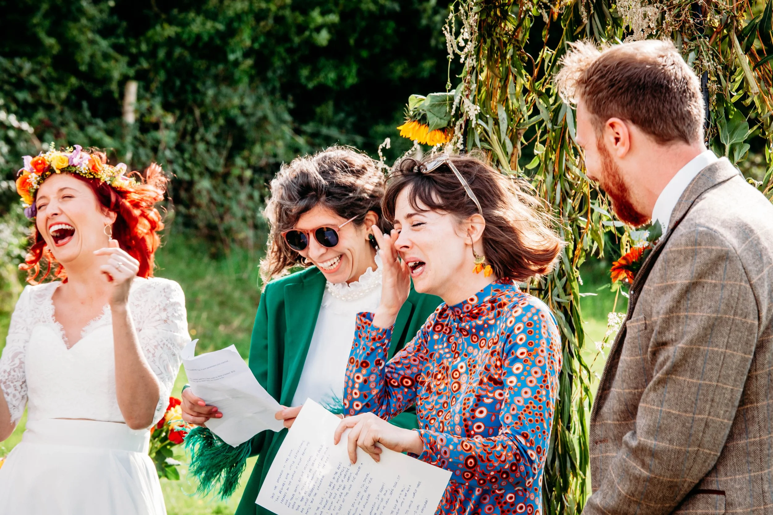 A bride wearing a colourful flower crown bursts into laughter with her wedding guests during a joyful outdoor wedding ceremony.