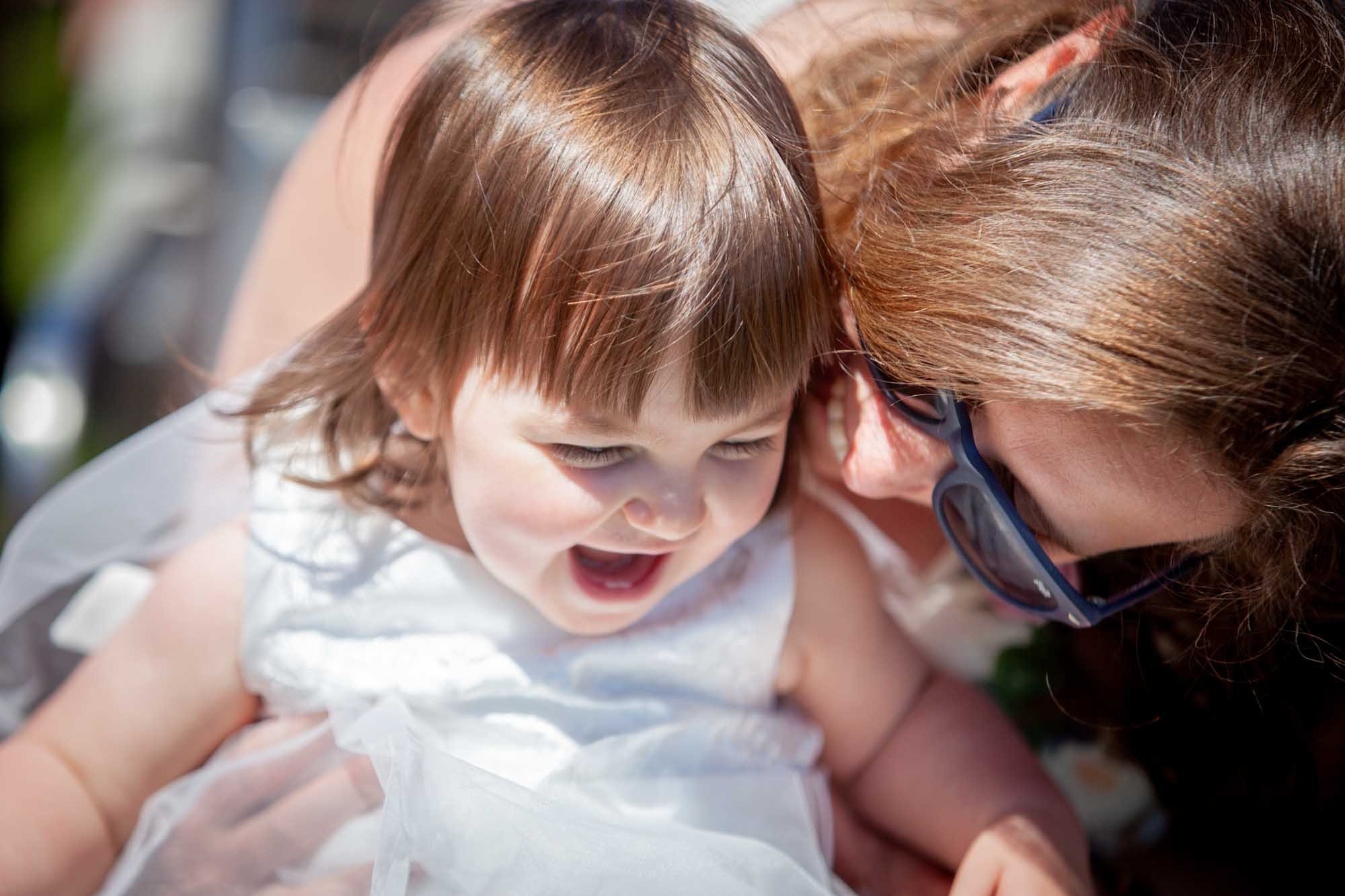 A little girl in a white dress laughs as a woman wearing sunglasses leans close to her during a wedding celebration.