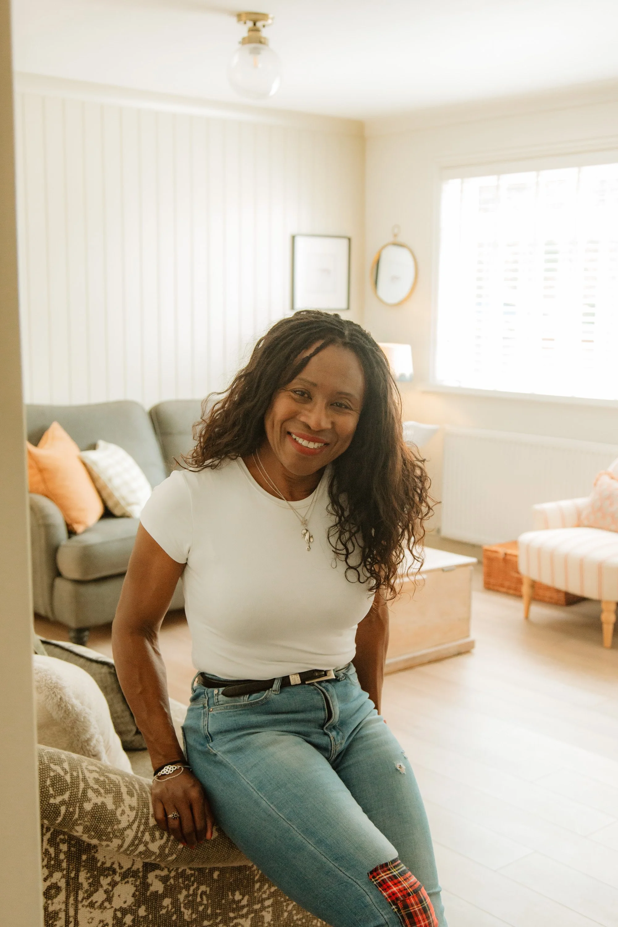 Confident woman smiling while seated on the arm of a sofa in a light-filled, cozy home interior
