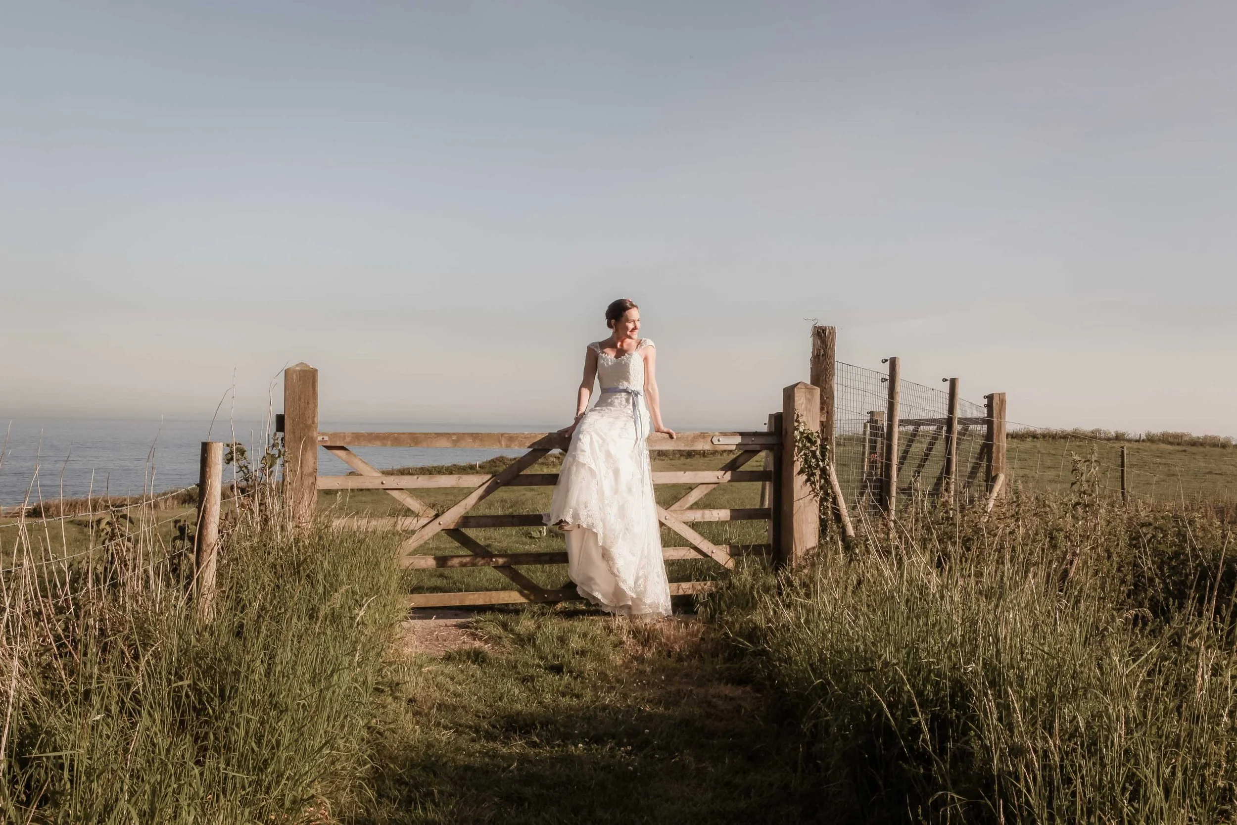 Bride beside a countryside gate overlooking the coast on a sunny Isle of Wight summer wedding day.