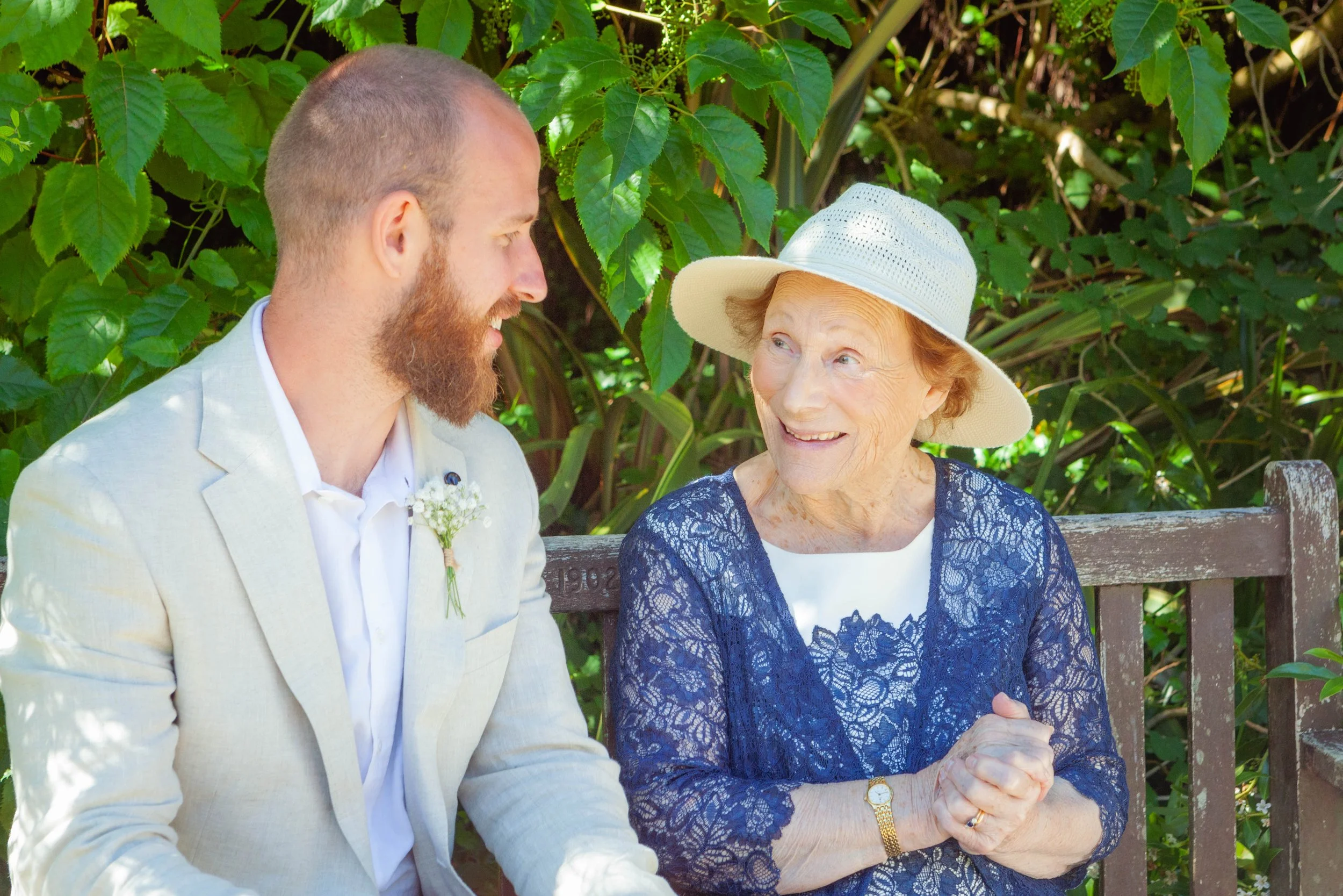 A groom in a light linen suit shares a warm smile with an elderly wedding guest wearing a blue lace dress and sun hat, seated together on a garden bench.