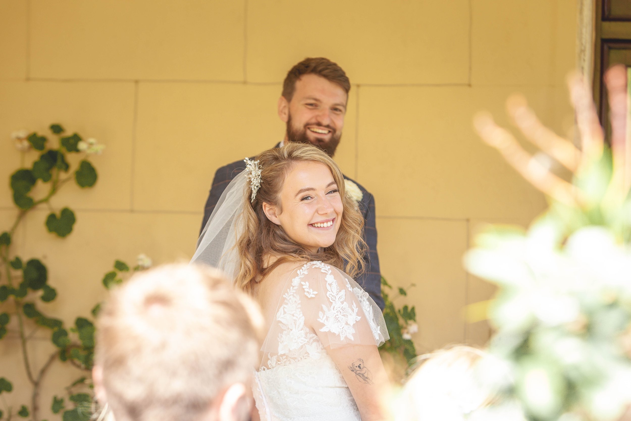 A smiling bride in a lace wedding dress looks back towards her guests, with her groom smiling behind her during an intimate wedding ceremony.