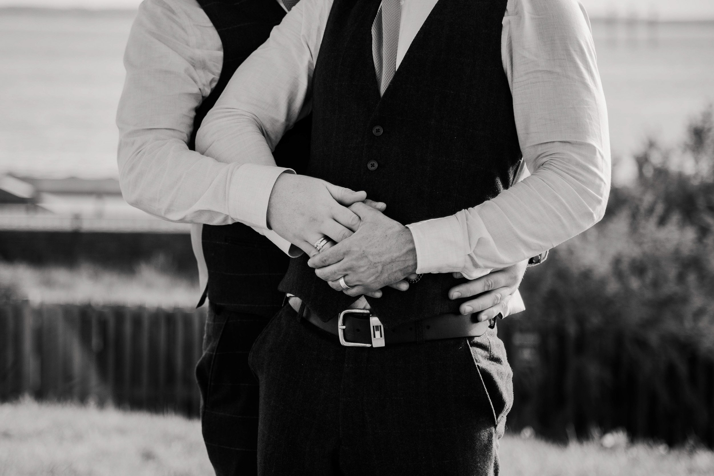 Close-up black-and-white wedding photograph of two grooms embracing, their hands resting together with wedding rings visible.