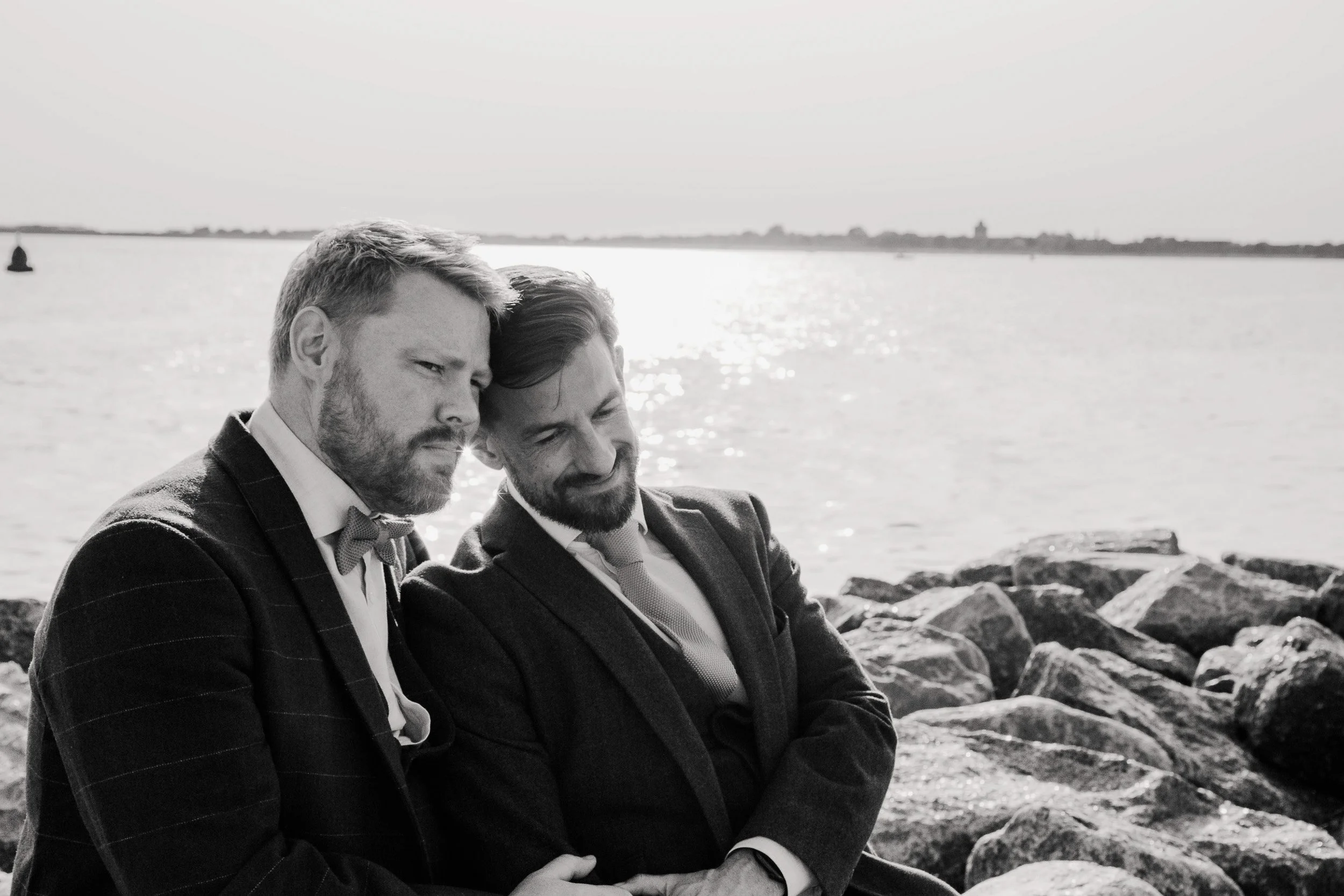 Two grooms rest their heads together in a quiet, intimate moment beside the sea, captured in black and white during their Isle of Wight wedding.