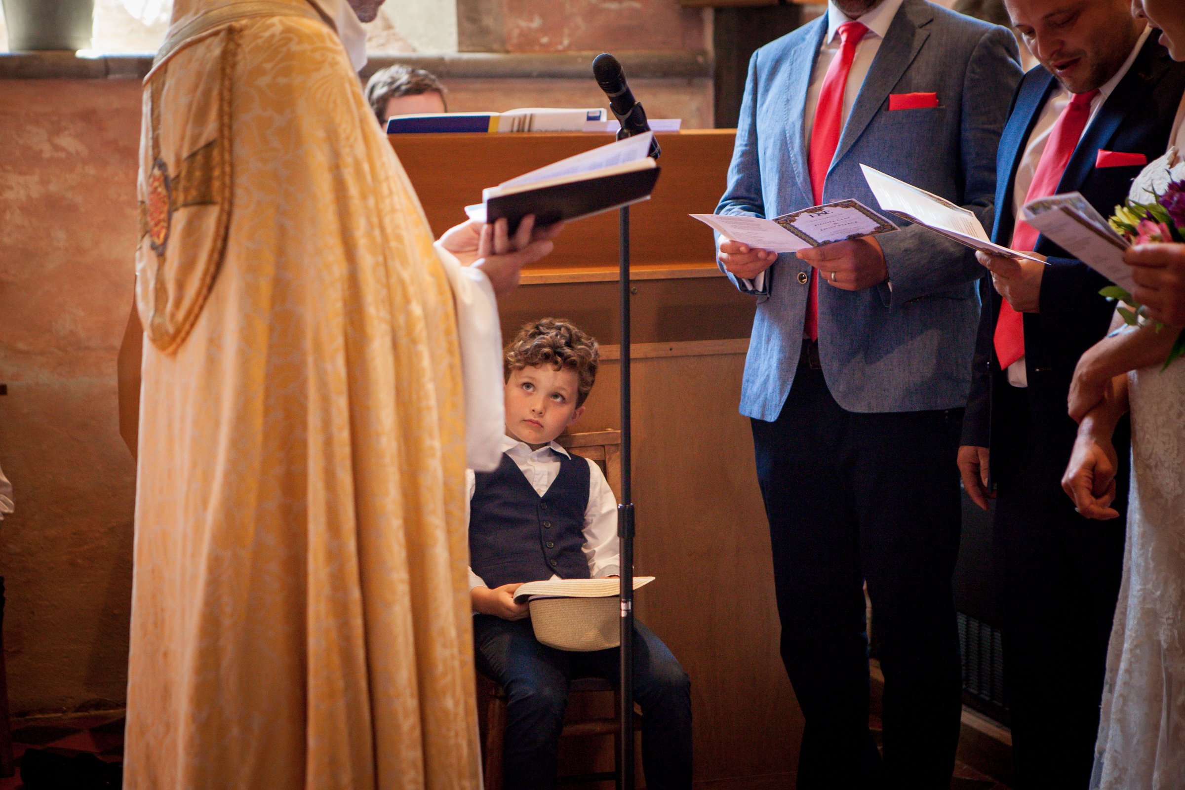 A young page boy in a navy waistcoat and bow tie looks up curiously during a church wedding ceremony as the adults read from their service booklets.
