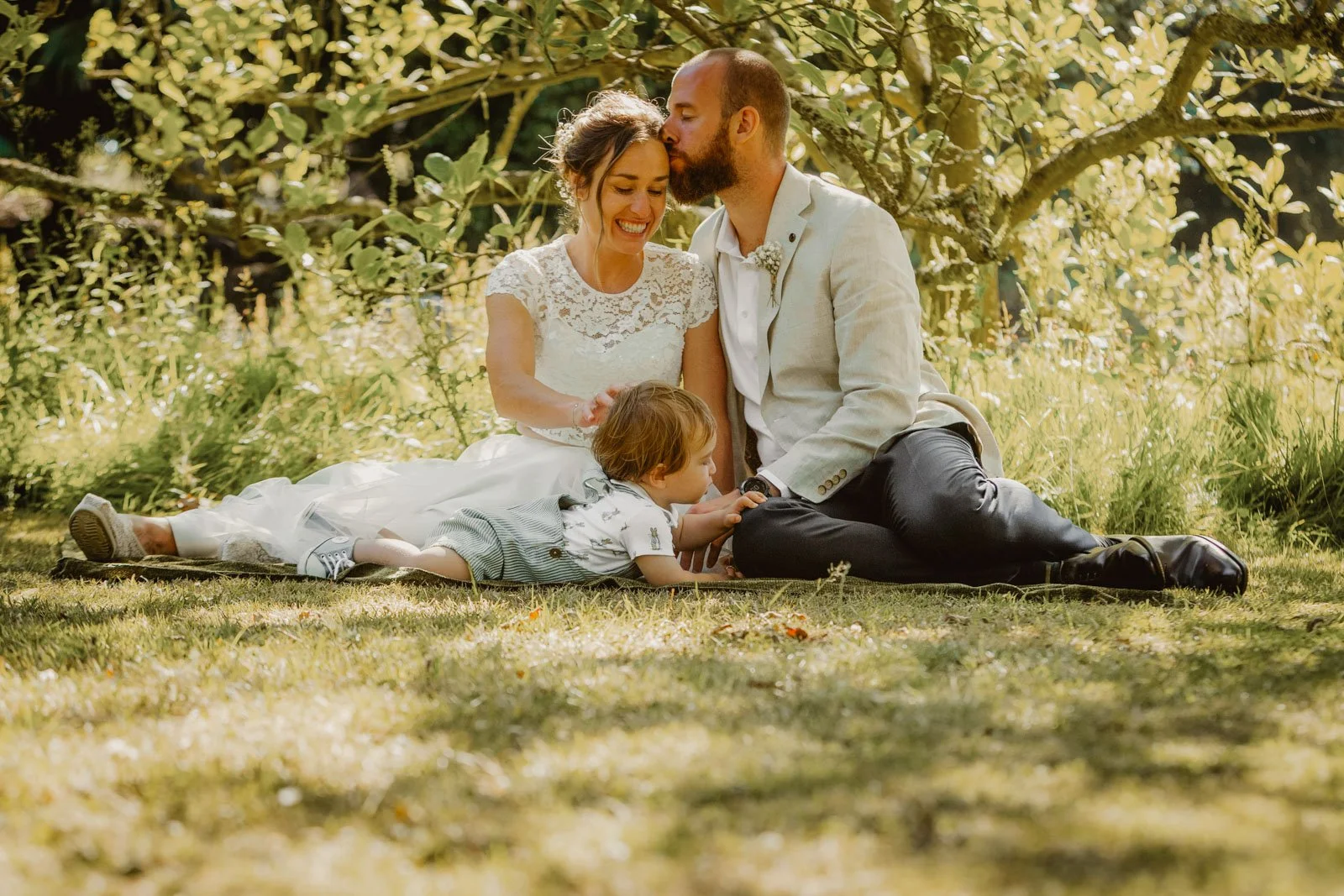 Bride and groom relaxing with their young child in the shade during a sunny summer wedding on the Isle of Wight.
