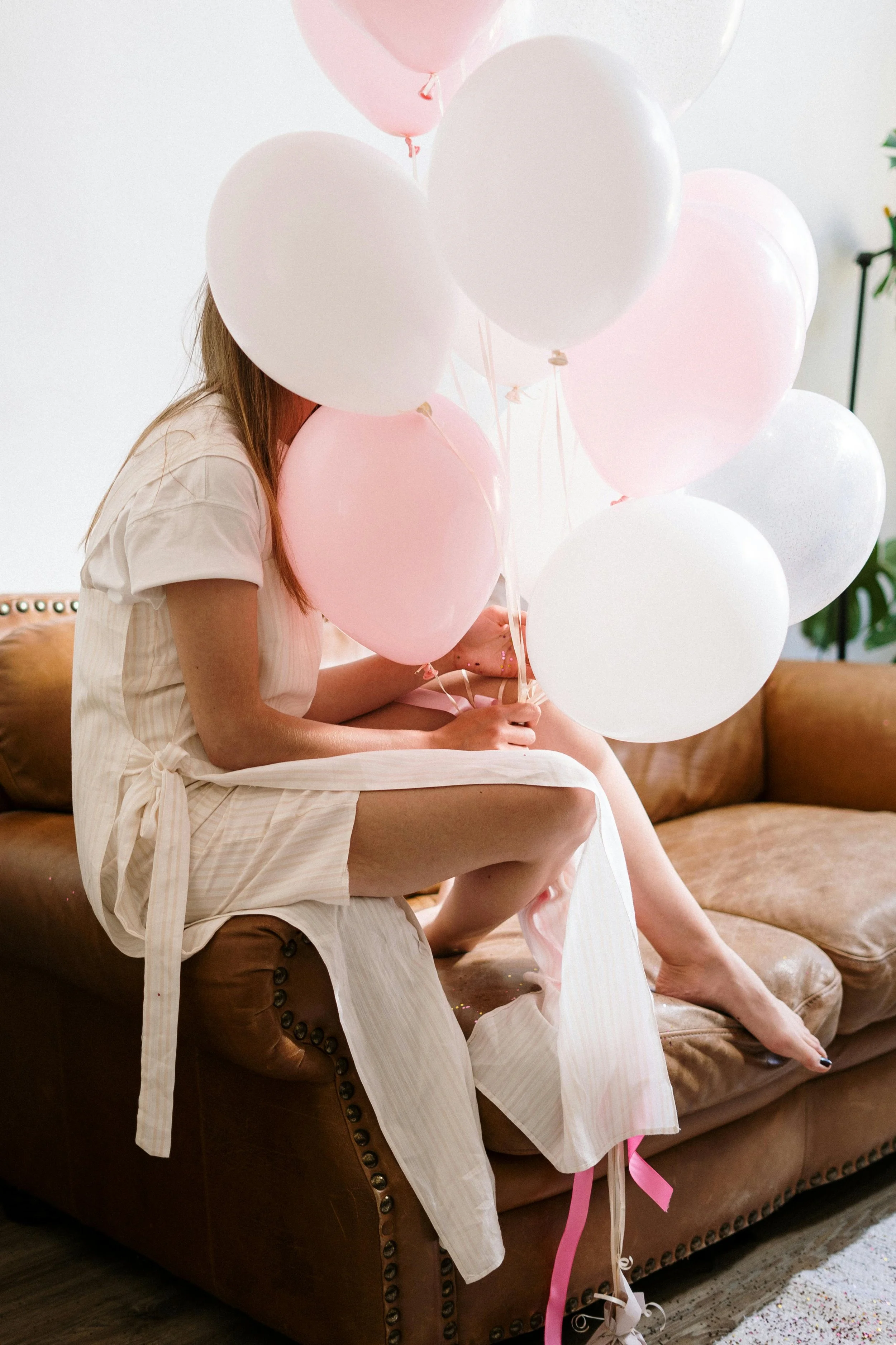 Person sitting on a brown leather couch holding pink and white balloons, with face obscured by the balloons.