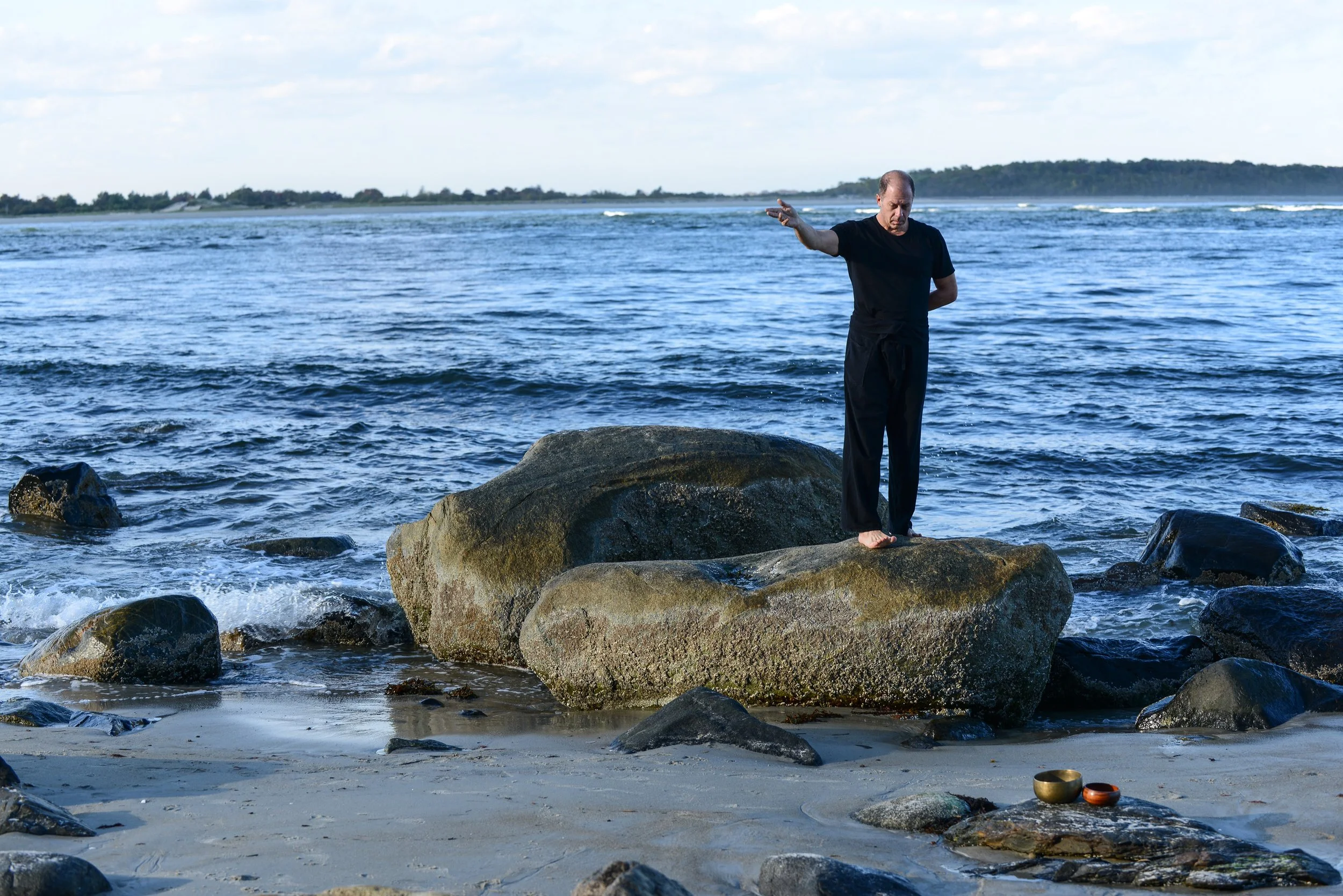 A man practicing yoga on a large rock by the ocean, with waves and sky in the background, and bowls placed on the sand.