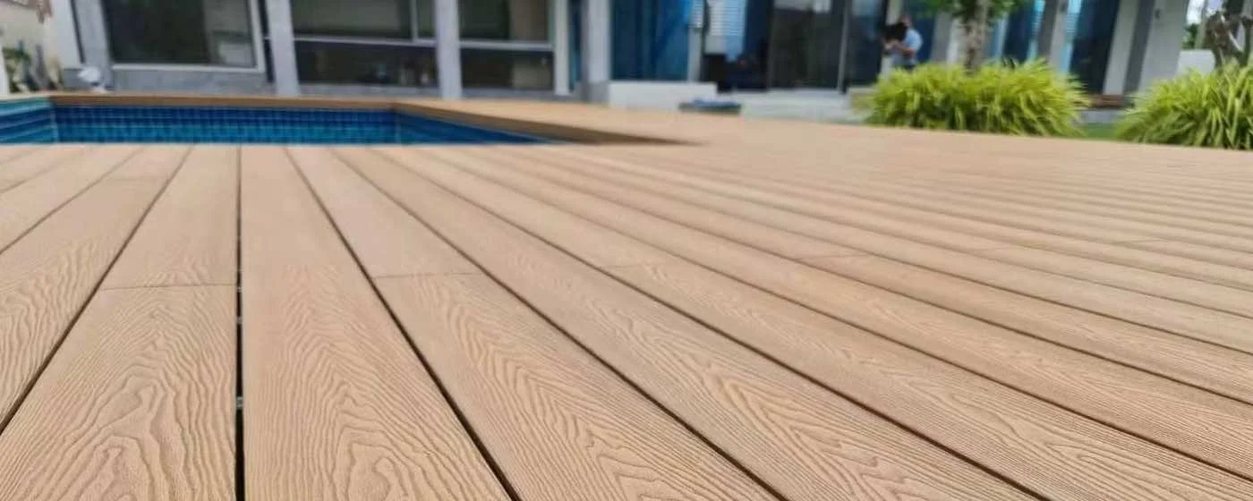 A view of a wooden pool deck with a swimming pool in the background, surrounded by a modern patio area and greenery.