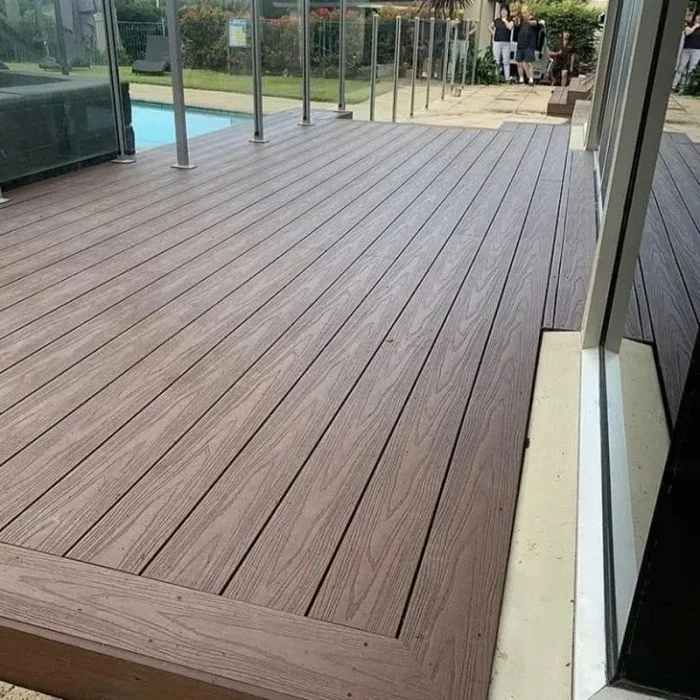 View of a modern outdoor wooden deck with glass railing, adjacent to a building with a sliding glass door, overlooking a pool and a grassy backyard with people in the background.