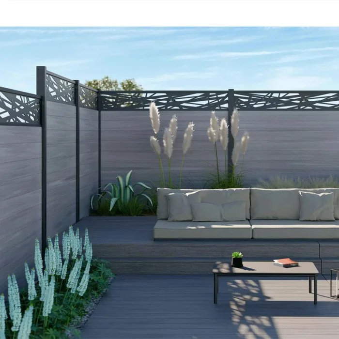 Outdoor patio with gray wooden decking, a gray sectional sofa with cushions, potted plants including pampas grass and other greenery, and a black coffee table with a small plant and a book, enclosed by gray privacy fencing.