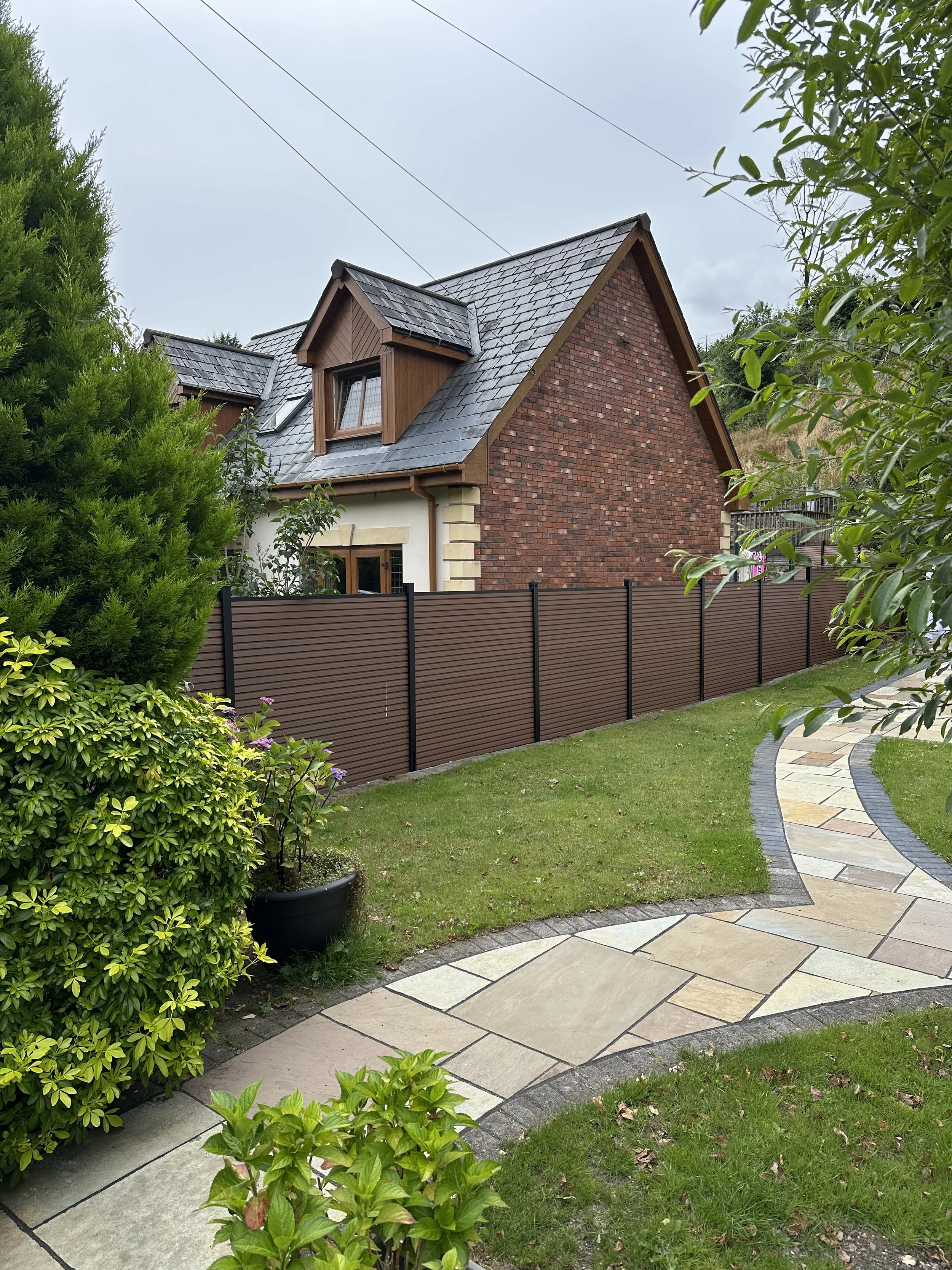 A house with a brick exterior and a slate roof, surrounded by a garden with a curved stone pathway, green bushes, and trees, enclosed by a brown fence.