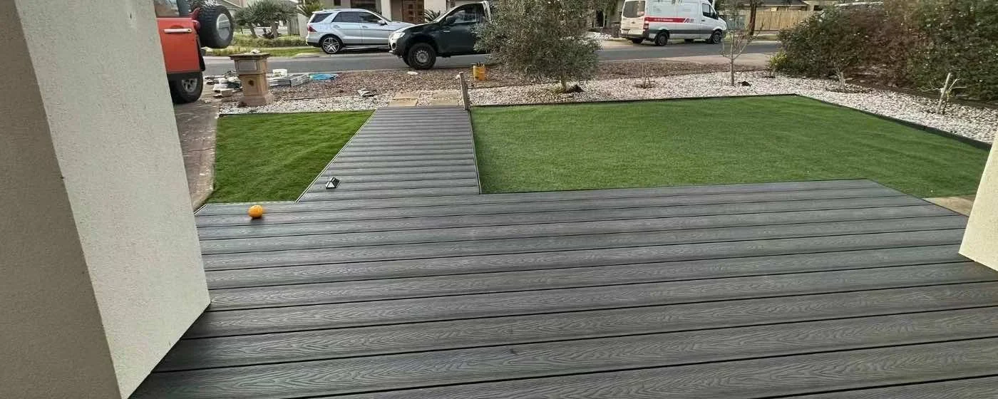 View from a porch overlooking a backyard with a deck, artificial grass, and desert landscaping. Parked cars are visible across the street.