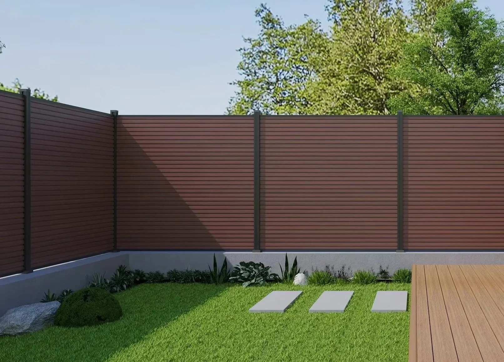 A backyard with a green lawn, three white stepping stones, a brown wooden deck, a row of various small plants, a large bush, and a brown fence with metal posts, with trees and a blue sky in the background.