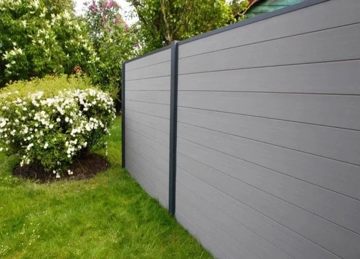 A gray vinyl privacy fence in a backyard, with green grass and flowering bushes nearby under a partly cloudy sky.