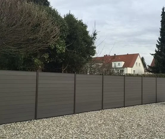 A tall gray privacy fence along a gravel path with trees and a house with a red roof in the background.