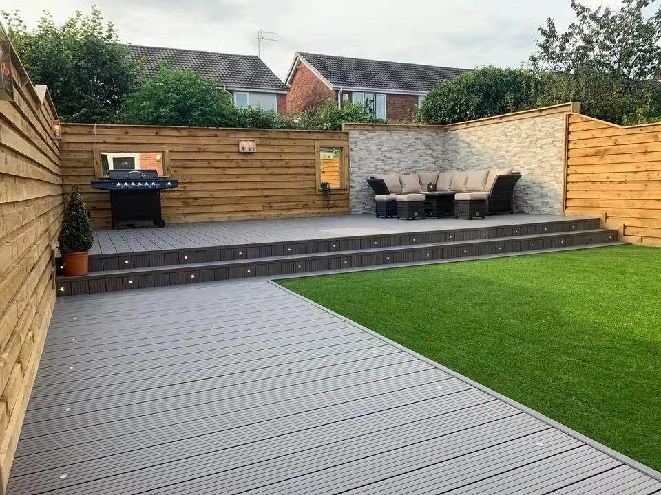 A modern backyard patio with wooden decking, outdoor furniture, a barbecue grill, and a small patch of artificial grass, enclosed by wooden fences.