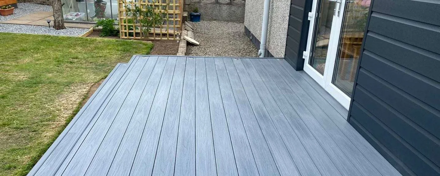 Newly installed gray wooden deck attached to a house with sliding glass doors, adjacent to a green lawn and a small garden area.