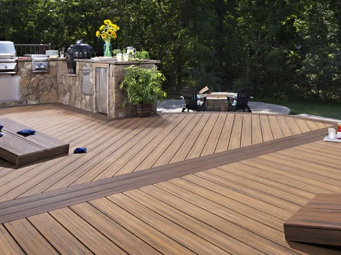 A backyard deck with wooden planks, a built-in outdoor kitchen with a stone facade, a table with four chairs, and a tree in the background.