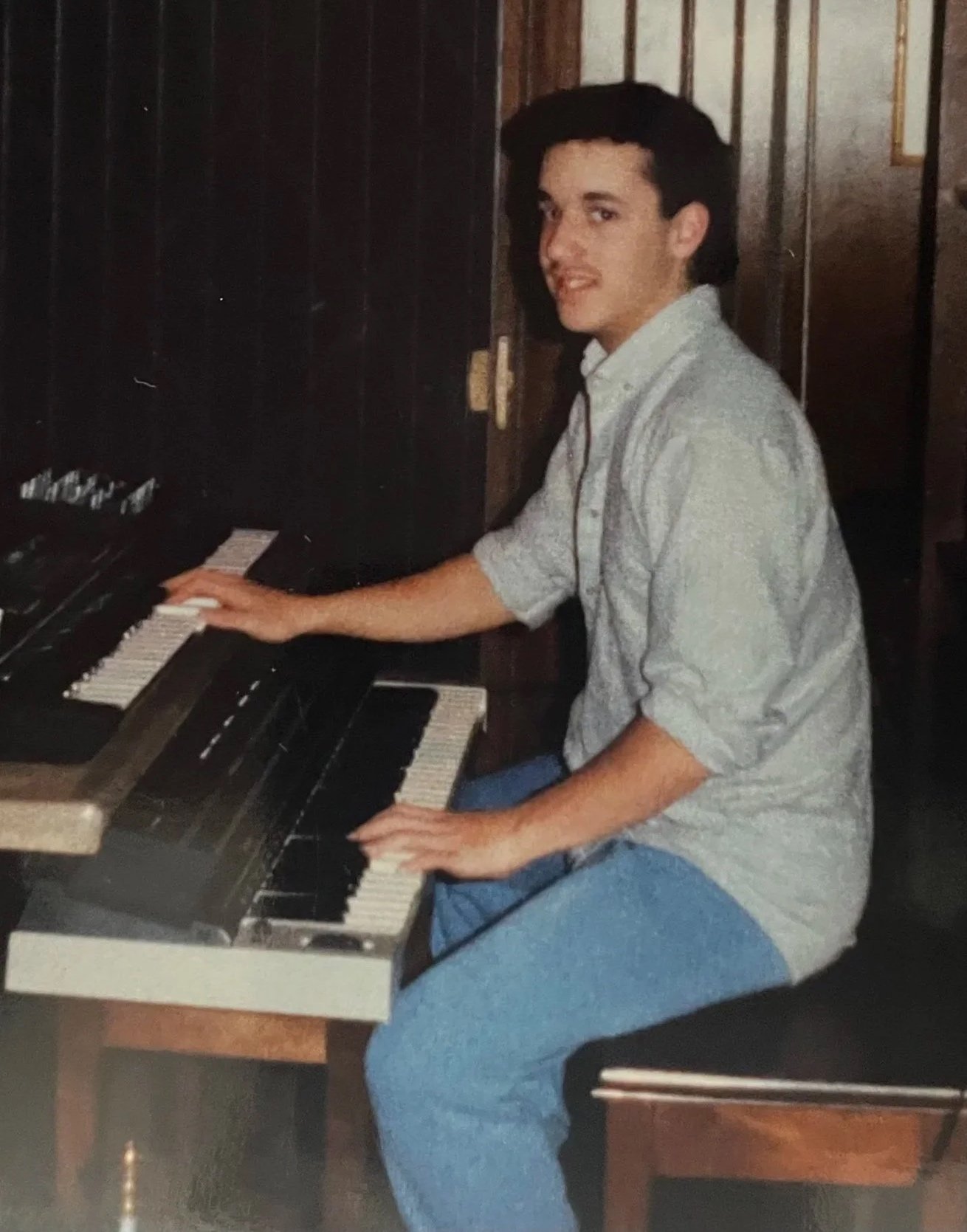 A young man playing a piano, sitting on a wooden bench, in a room with wood-paneled walls, wearing a light-colored button-up shirt with rolled-up sleeves and blue pants.