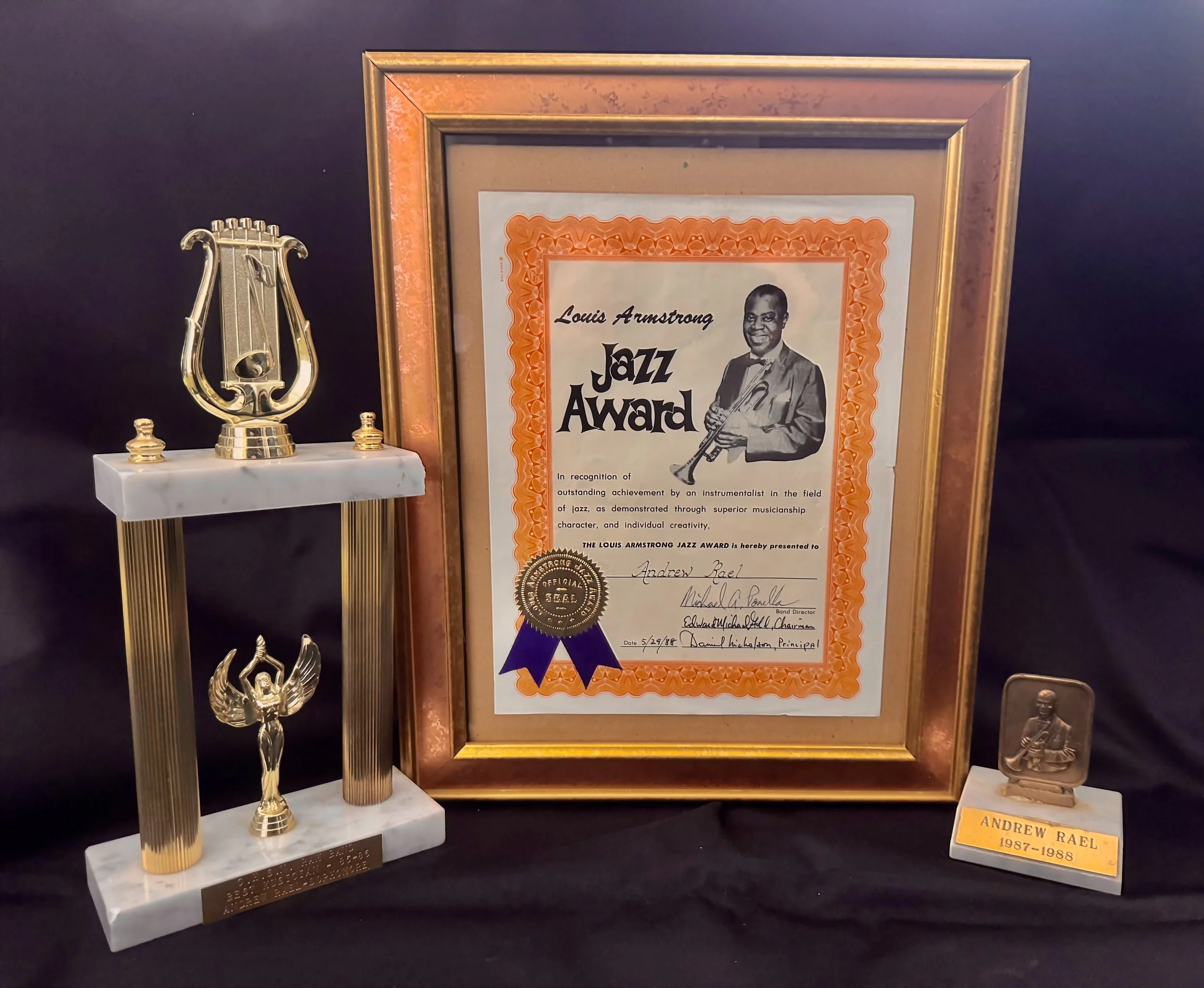 Awards display including a framed Louis Armstrong Jazz Award certificate, a gold and white trophy with a lyre and angel figure, second Louis Armstrong award, best jazz musician and best overall musician