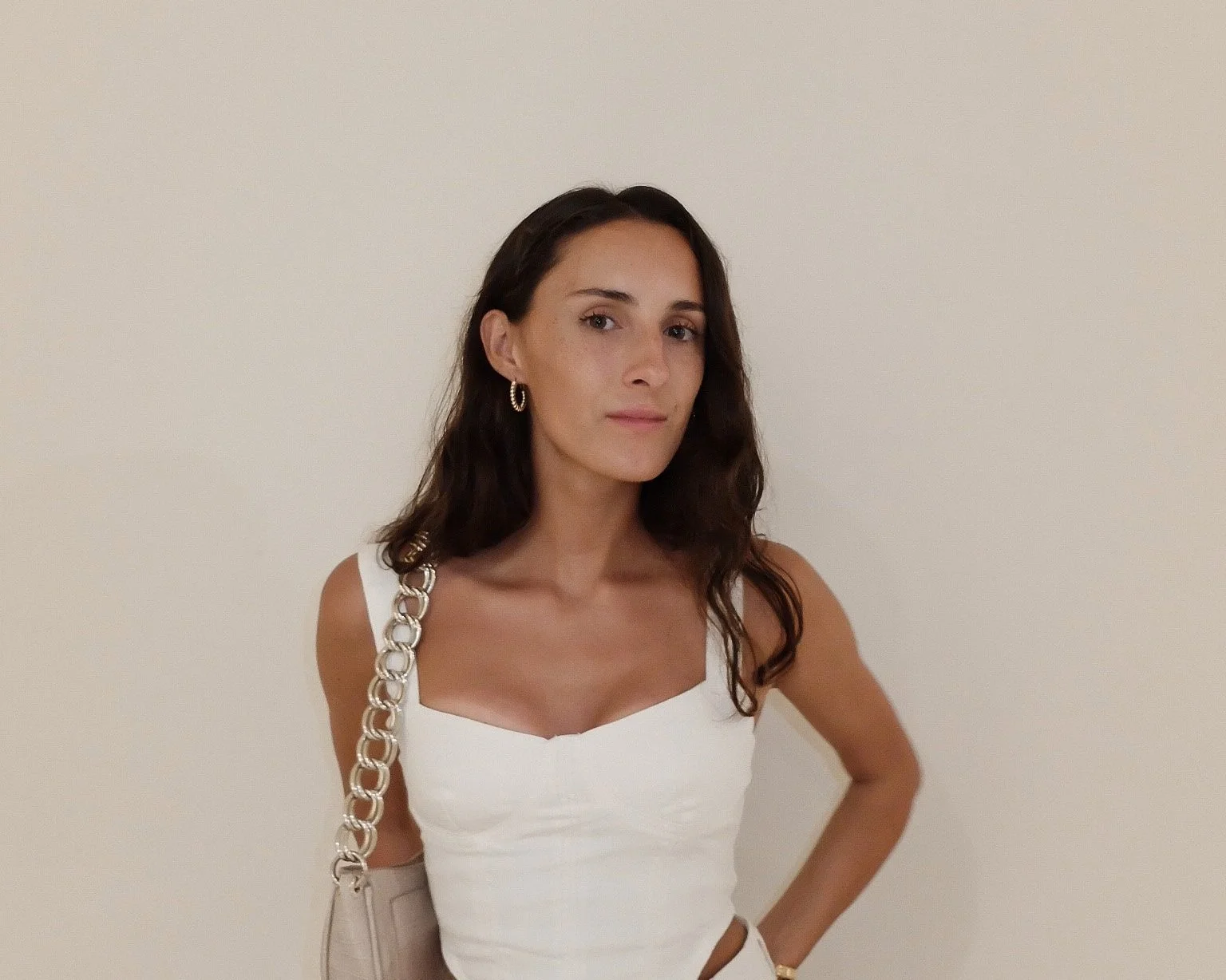 A woman with long dark hair wearing a white sleeveless top, gold hoop earrings, and a beige handbag with chain straps, standing against a plain beige wall.